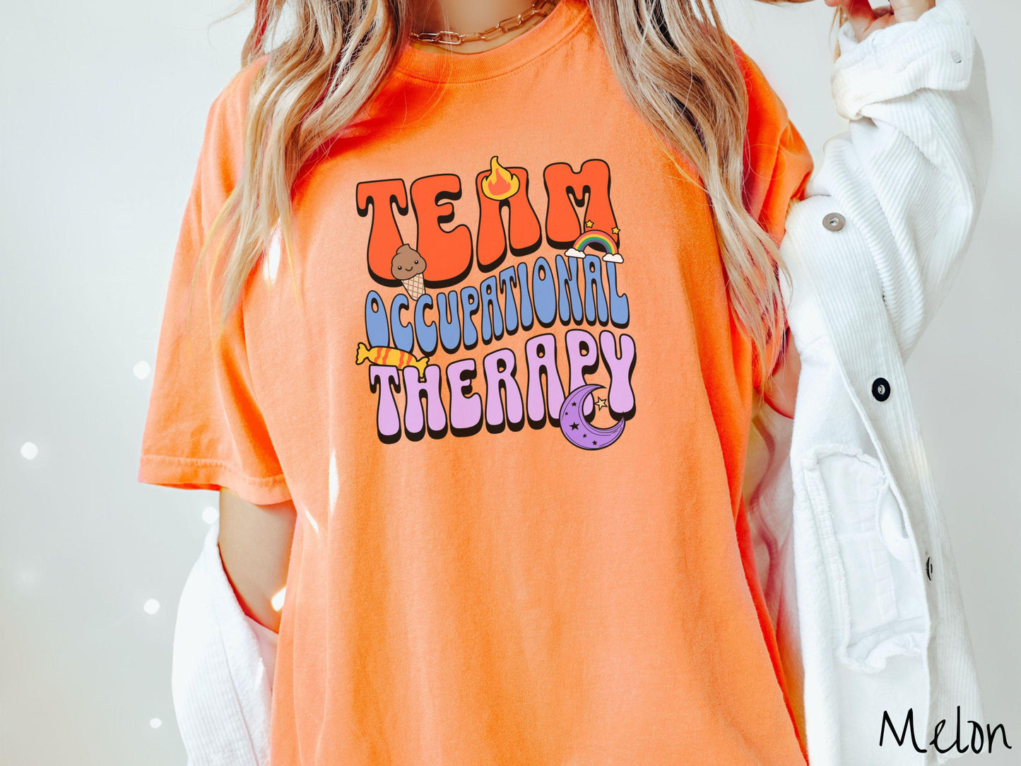 A vintage, melon colored Comfort Colors t-shirt with the text "Team Occupational Therapy" surrounded by playful elements like a chocolate ice cream cone, a blazing flame, a colorful rainbow, a piece of candy, and a serene moon.