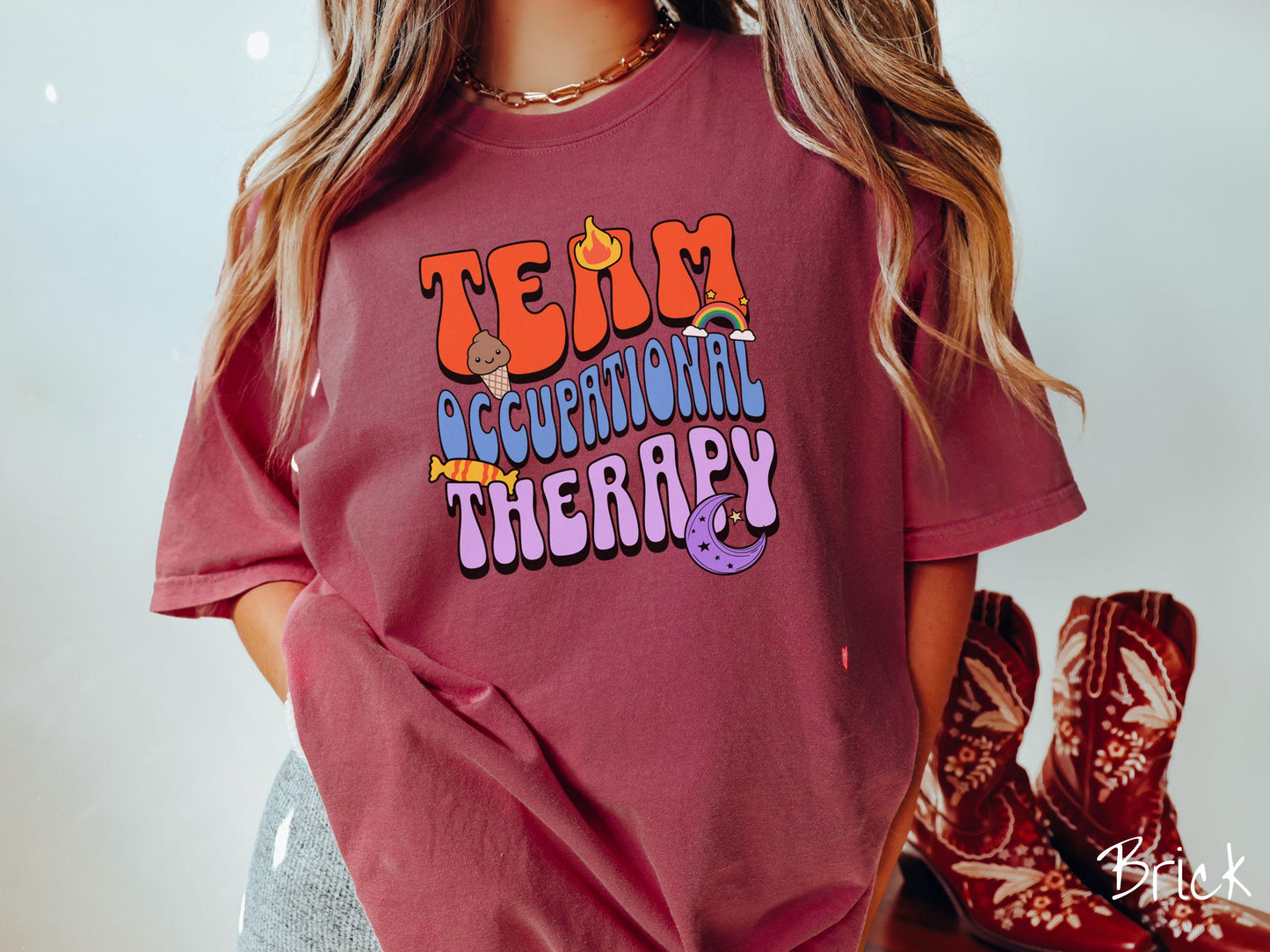 A vintage, brick colored Comfort Colors t-shirt with the text "Team Occupational Therapy" surrounded by playful elements like a chocolate ice cream cone, a blazing flame, a colorful rainbow, a piece of candy, and a serene moon.