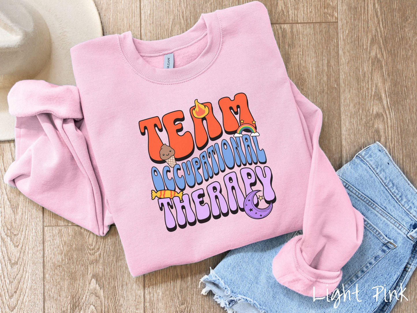A vintage, light pink colored Gildan sweatshirt with the text "Team Occupational Therapy" surrounded by playful elements like a chocolate ice cream cone, a blazing flame, a colorful rainbow, a piece of candy, and a serene moon.