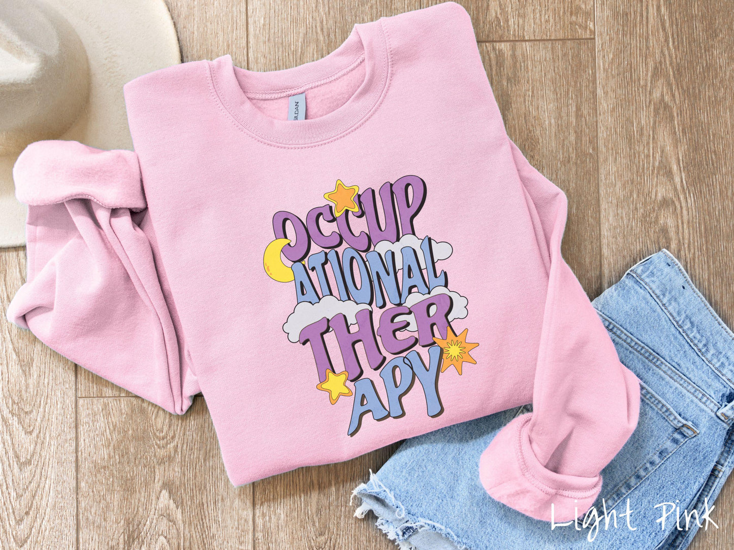A vintage, light pink colored Gildan sweatshirt with the text Occupational Therapy vertically with clouds, the yellow moon, and bright yellow and orange stars mixed in.