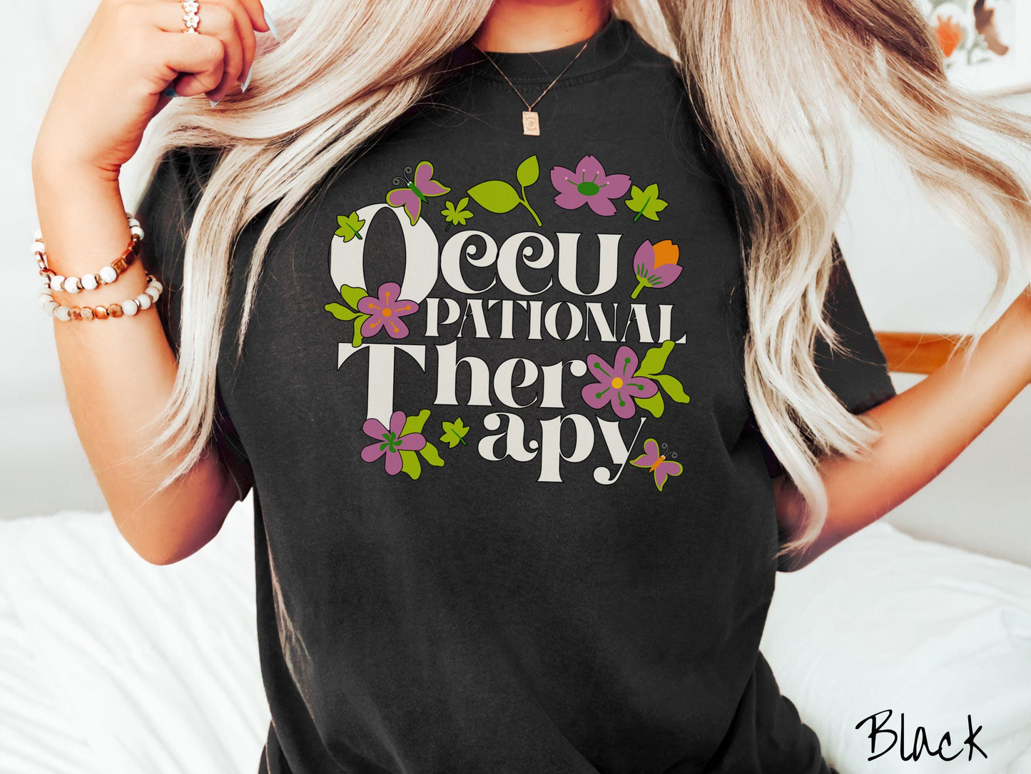 A vintage, black colored Comfort Colors t-shirt with the text Occupational Therapy and featuring green leaves, purple and orange flowers, and colorful butterflies in the background.
