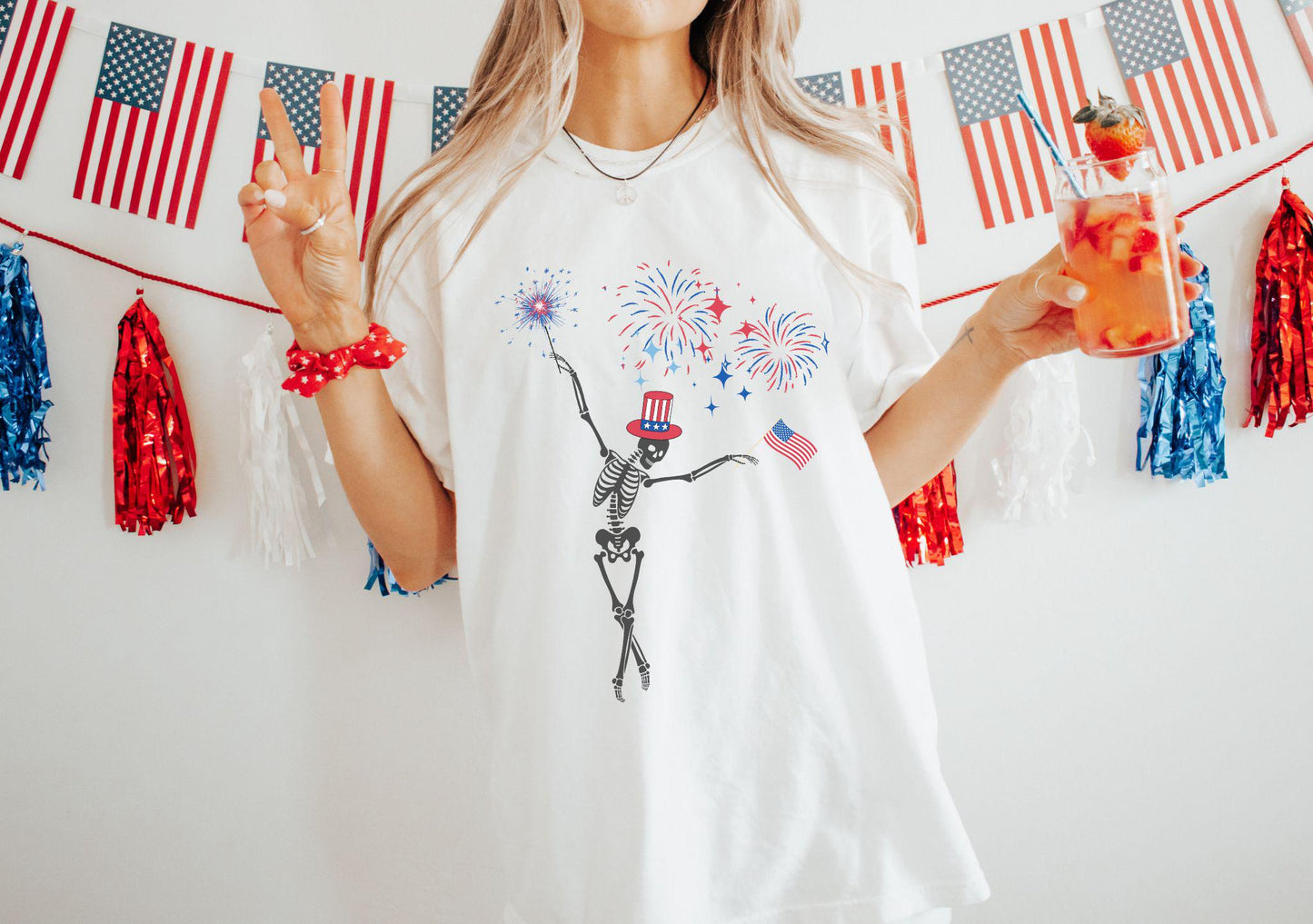 This Comfort Colors t-shirt features a skeleton wearing a red, white, and blue top hat, dancing in front of exploding fireworks while holding a sparkler firework and an American flag.