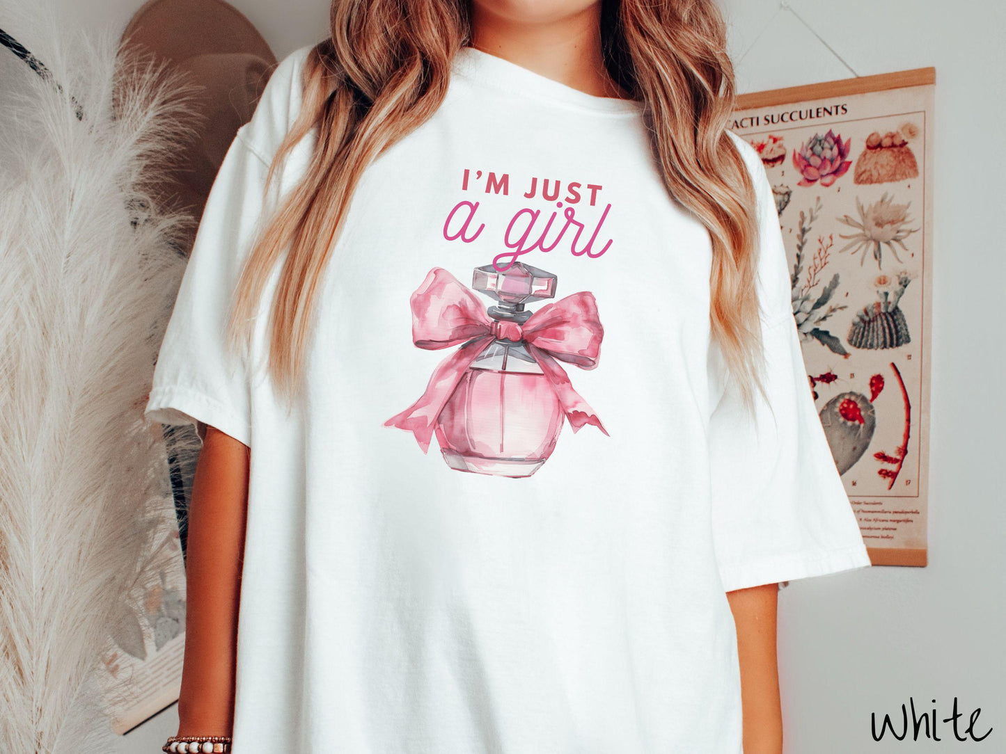 A stylish Comfort Colors T-shirt featuring the phrase "I'm Just a Girl" in pink font above a bottle of pink perfume tied with a pink bow. This tee exudes a feminine and trendy vibe, perfect for those who appreciate unique fashion statements.