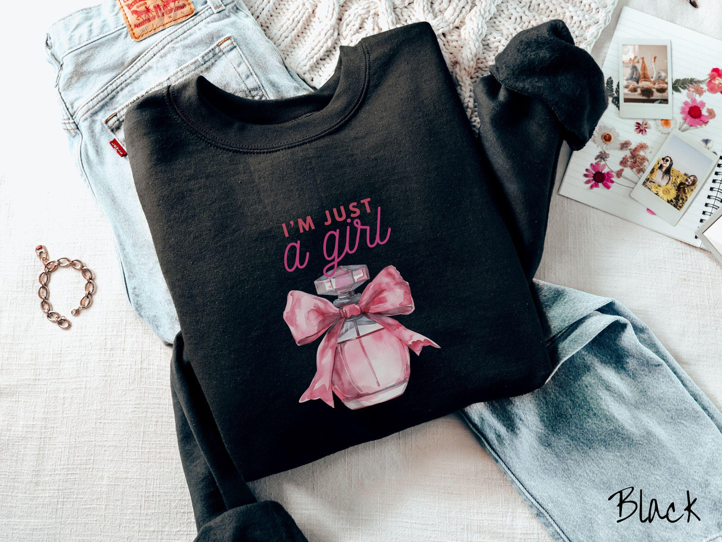 A cozy Gildan sweatshirt featuring the phrase "I'm Just a Girl" in pink font above a bottle of pink perfume tied with a pink bow. Perfect for those colder months, this sweatshirt provides warmth and comfort with its classic fit and crew neckline.