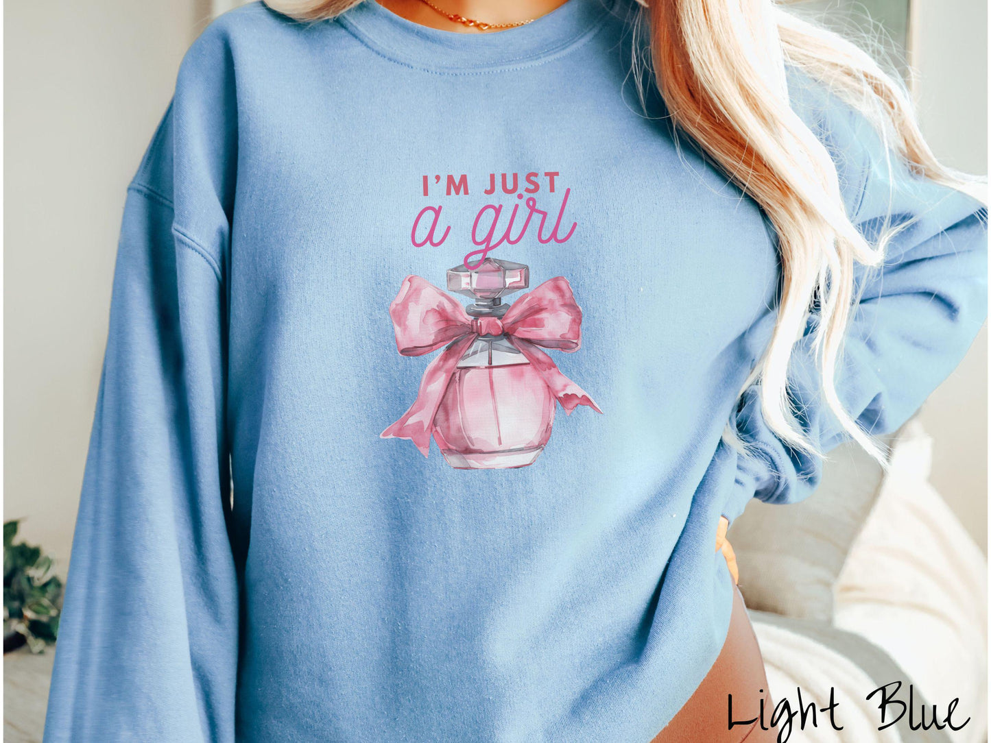 A cozy Gildan sweatshirt featuring the phrase "I'm Just a Girl" in pink font above a bottle of pink perfume tied with a pink bow. Perfect for those colder months, this sweatshirt provides warmth and comfort with its classic fit and crew neckline.