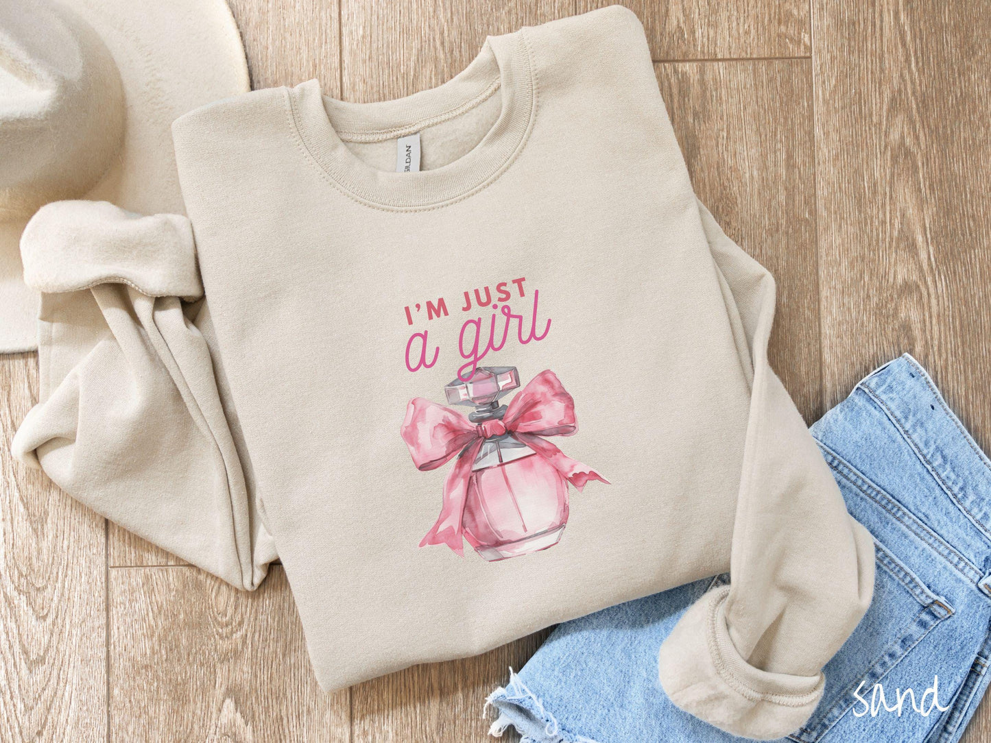 A cozy Gildan sweatshirt featuring the phrase "I'm Just a Girl" in pink font above a bottle of pink perfume tied with a pink bow. Perfect for those colder months, this sweatshirt provides warmth and comfort with its classic fit and crew neckline.