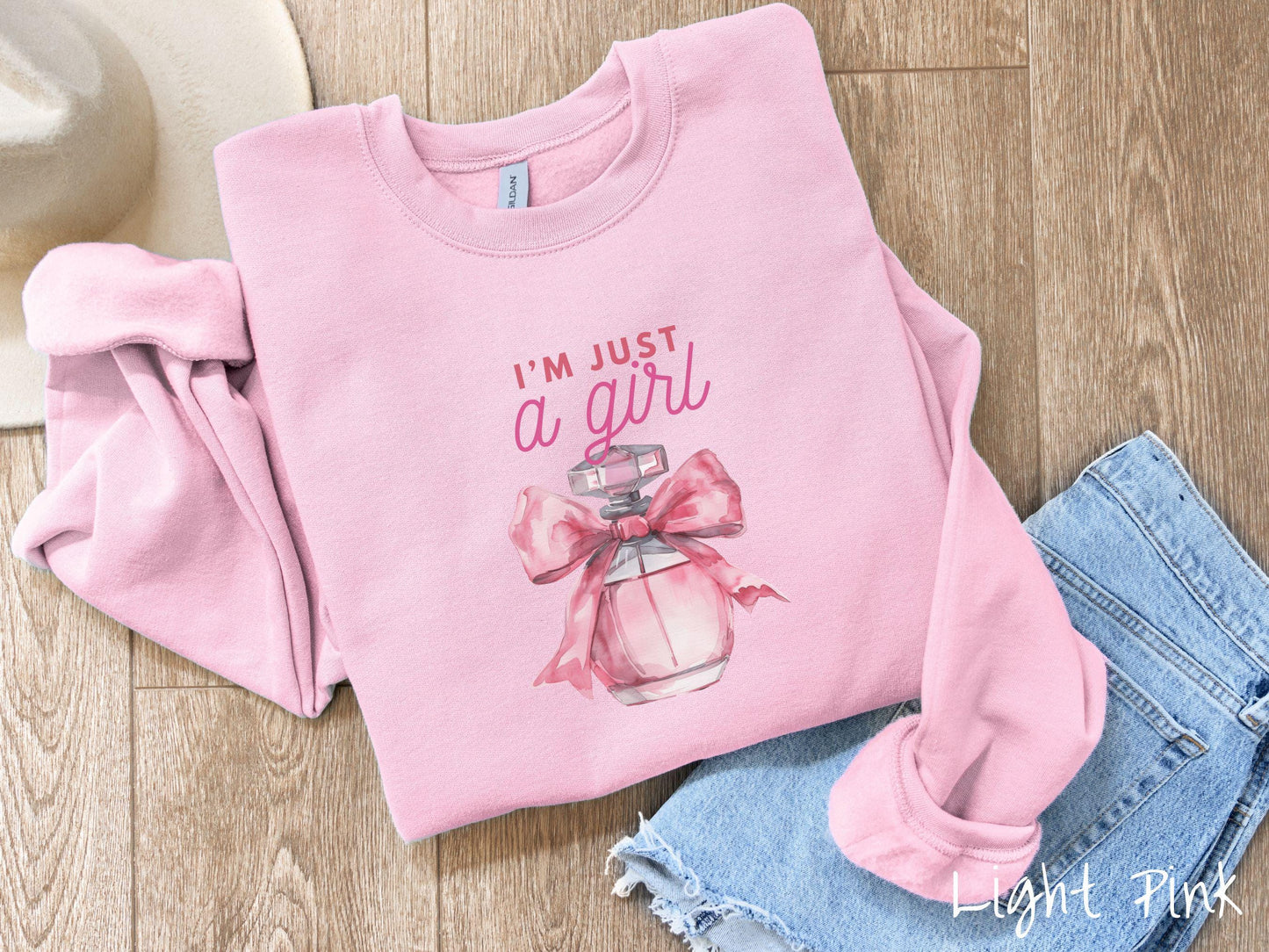 A cozy Gildan sweatshirt featuring the phrase "I'm Just a Girl" in pink font above a bottle of pink perfume tied with a pink bow. Perfect for those colder months, this sweatshirt provides warmth and comfort with its classic fit and crew neckline.