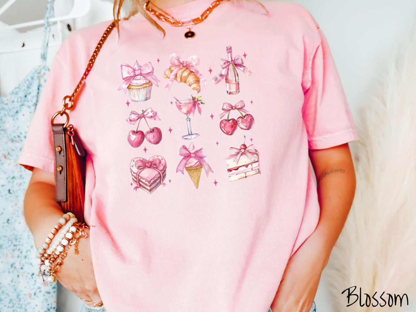 A stylish coquette Comfort Colors T-shirt featuring a grid of assorted treats with pink ribbons like a cupcake, a croissant, a champaigne bottle, cherries, a pink martini, a pink cake, and a pink ince cream cone, along with twinkling pink stars.