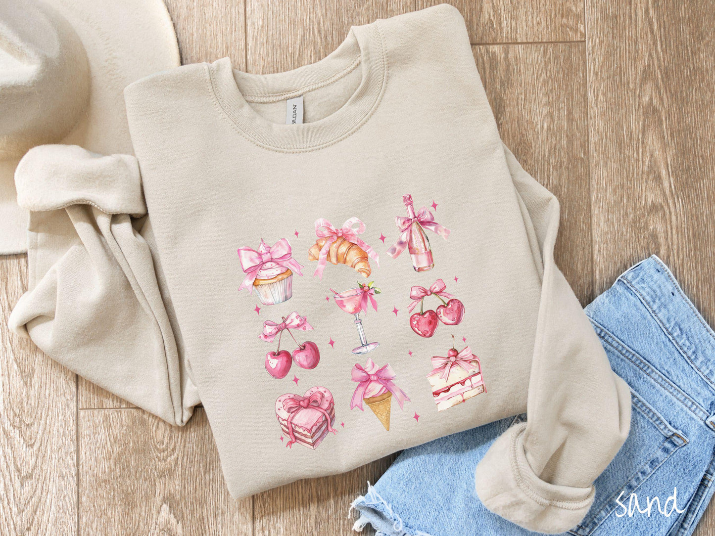 A stylish coquette Gildan sweatshirt featuring a grid of assorted treats with pink ribbons like a cupcake, a croissant, a champaigne bottle, cherries, a pink martini, a pink cake, and a pink ince cream cone, along with twinkling pink stars.
