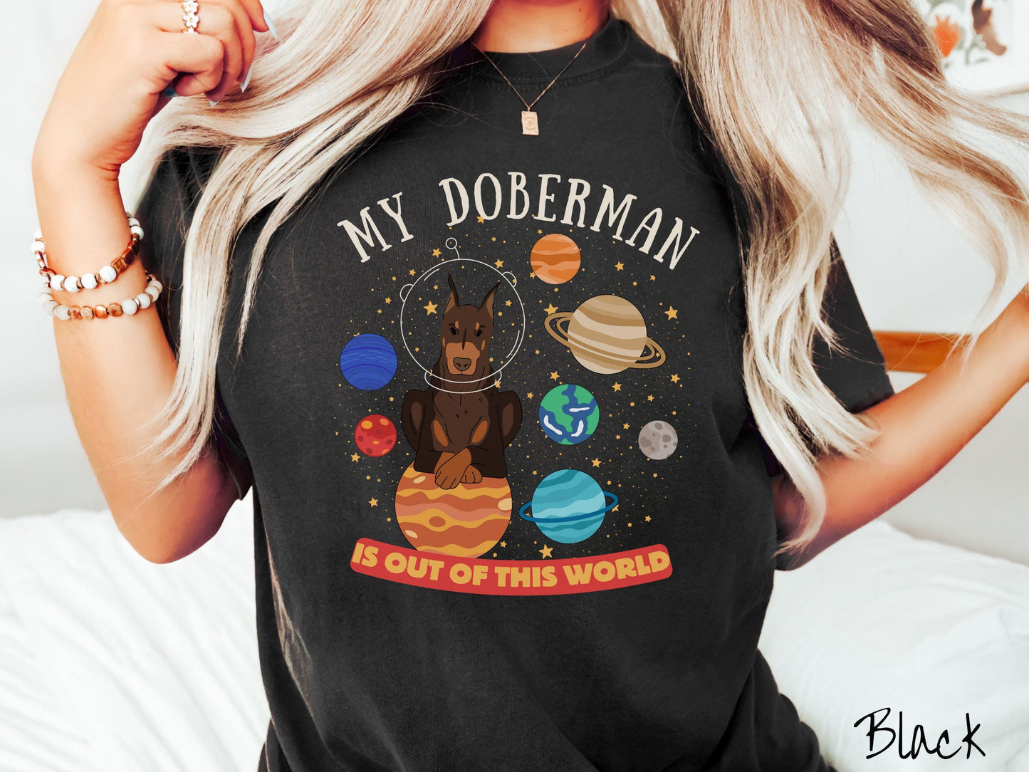 A vintage Comfort Colors t-shirt featuring the text My Doberman is Out of This World. The design showcases a Doberman sitting proudly atop Jupiter, surrounded by a vibrant solar system filled with colorful planets and twinkling stars.