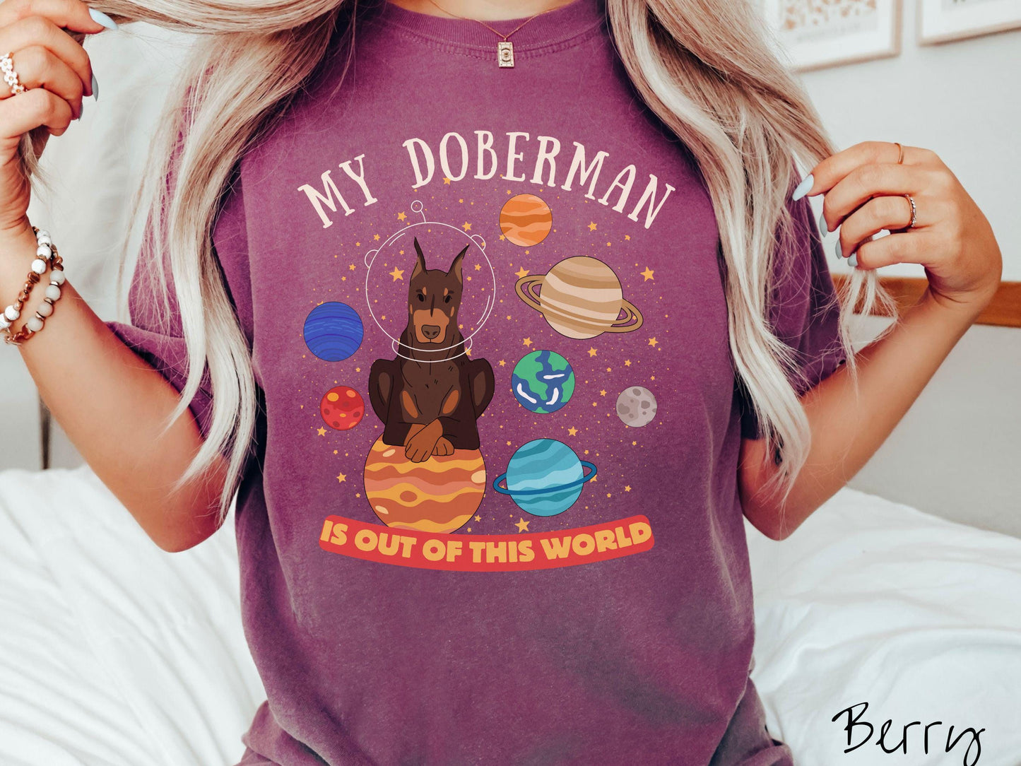 A vintage Comfort Colors t-shirt featuring the text My Doberman is Out of This World. The design showcases a Doberman sitting proudly atop Jupiter, surrounded by a vibrant solar system filled with colorful planets and twinkling stars.