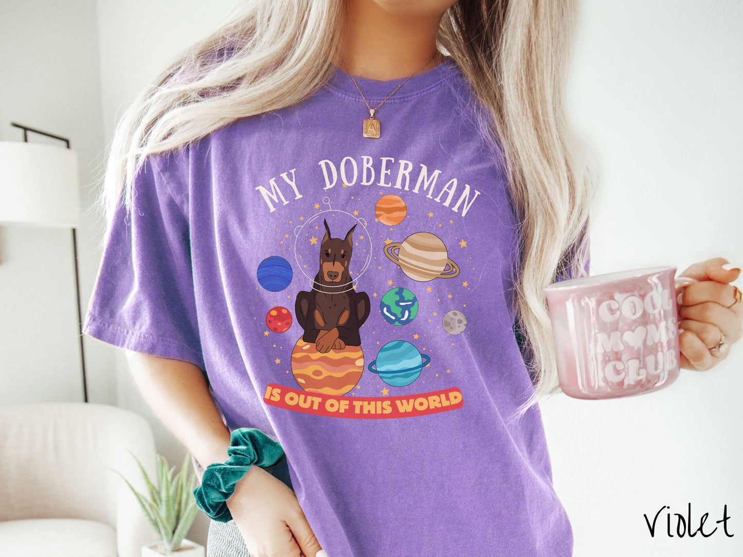 A vintage Comfort Colors t-shirt featuring the text My Doberman is Out of This World. The design showcases a Doberman sitting proudly atop Jupiter, surrounded by a vibrant solar system filled with colorful planets and twinkling stars.