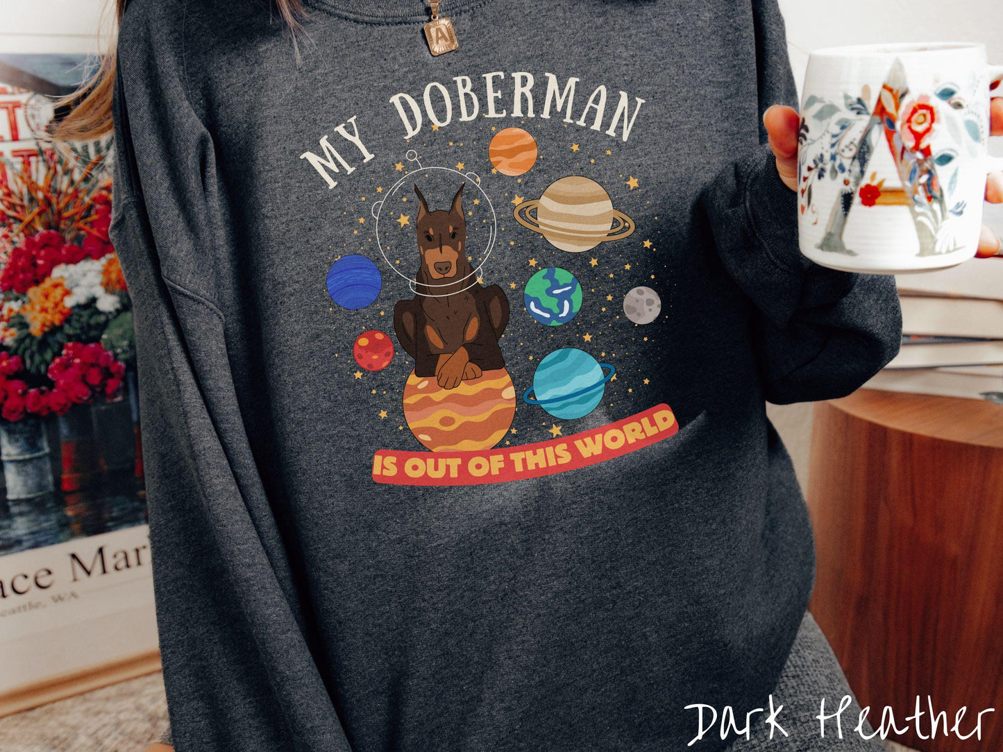 A vintage Gildan sweatshirt featuring the text My Doberman is Out of This World. The design showcases a Doberman sitting proudly atop Jupiter, surrounded by a vibrant solar system filled with colorful planets and twinkling stars.
