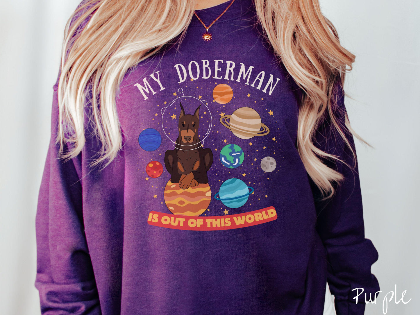 A vintage Gildan sweatshirt featuring the text My Doberman is Out of This World. The design showcases a Doberman sitting proudly atop Jupiter, surrounded by a vibrant solar system filled with colorful planets and twinkling stars.
