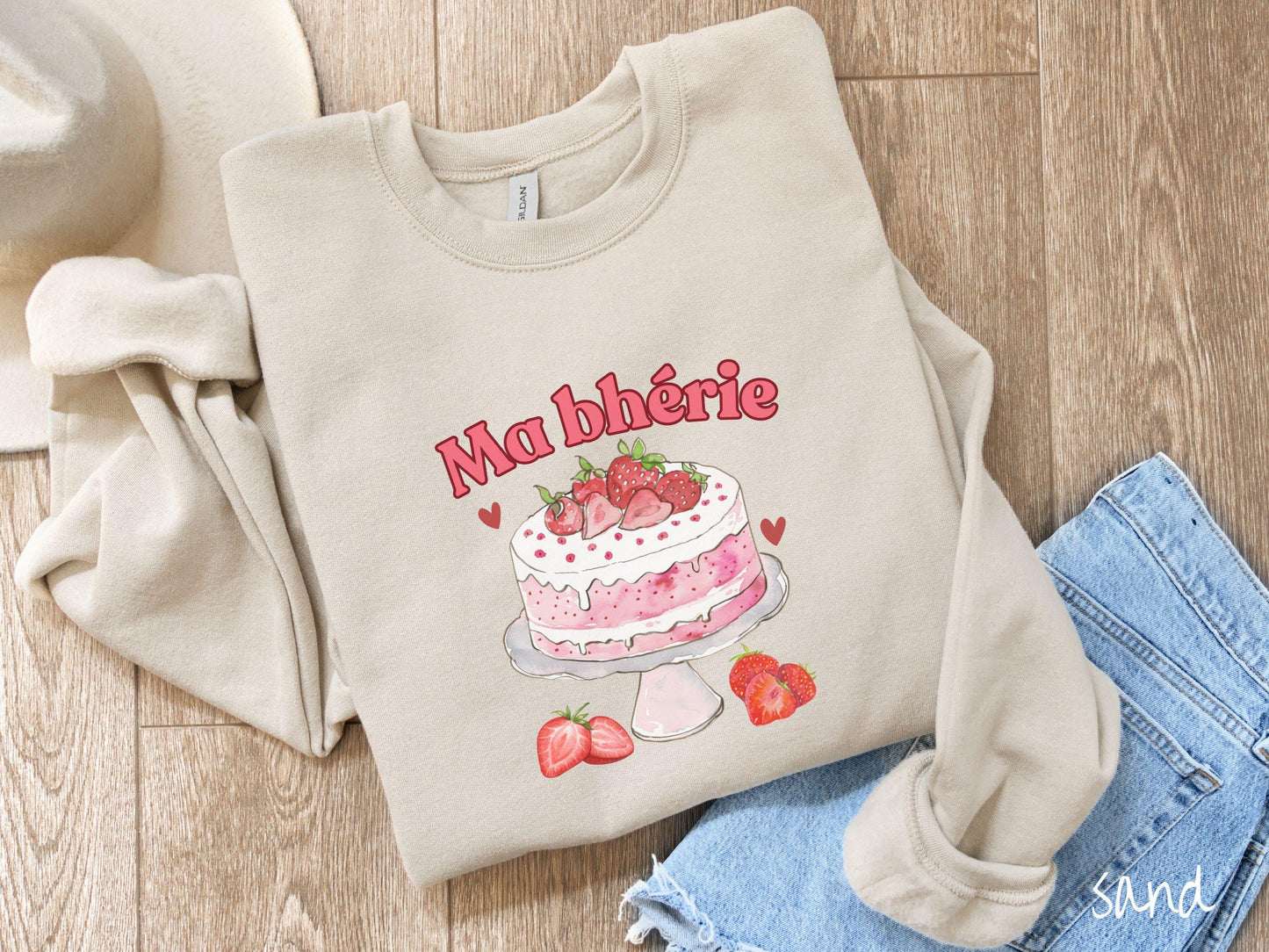 A Gildan sweatshirt featuring the phrase "Ma Bherie" is printed in bold red font above a charming illustration of a red-and-white strawberry cake displayed on a plate, topped with fresh strawberries and surrounded by even more berries.