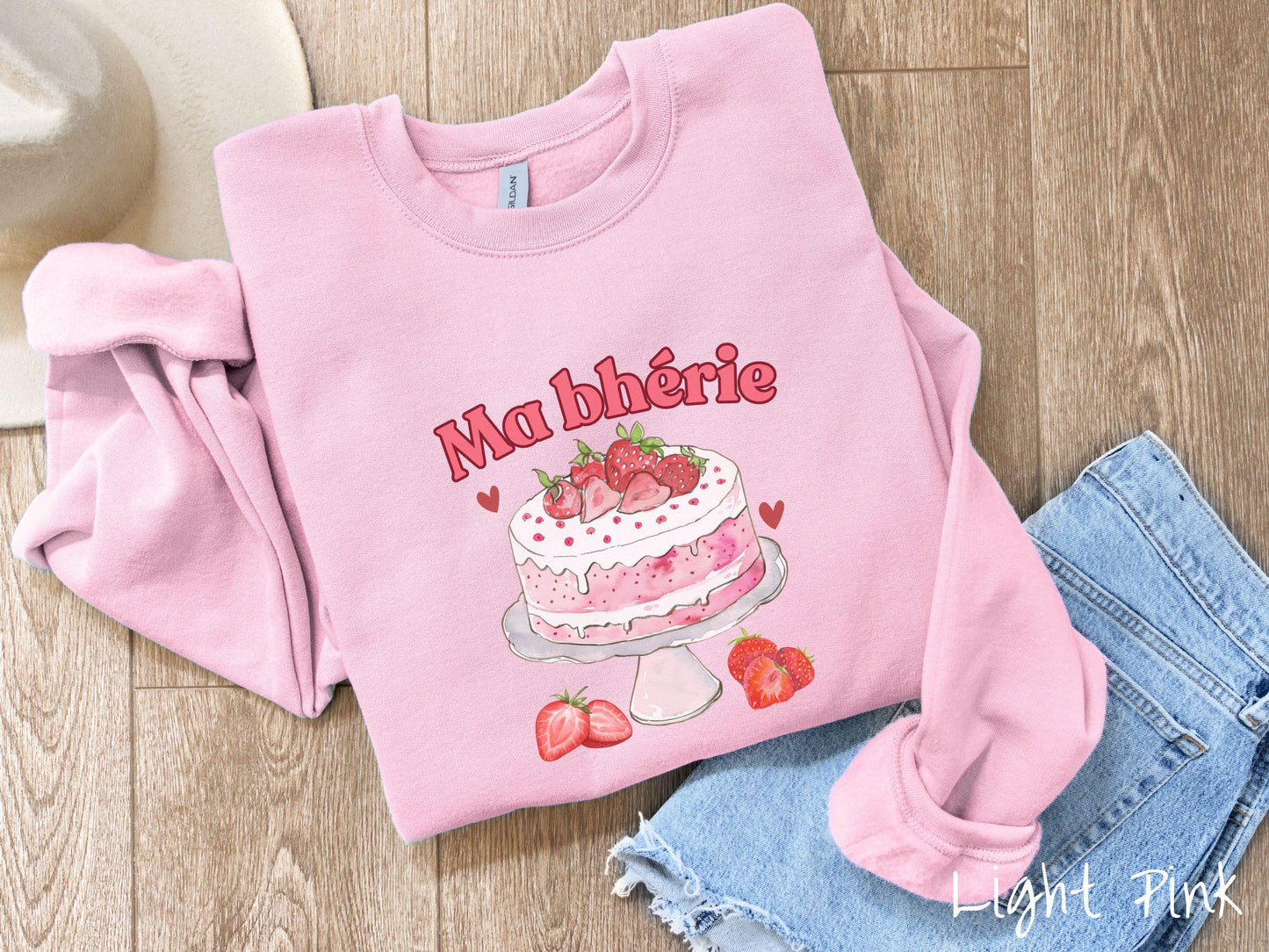 A Gildan sweatshirt featuring the phrase "Ma Bherie" is printed in bold red font above a charming illustration of a red-and-white strawberry cake displayed on a plate, topped with fresh strawberries and surrounded by even more berries.