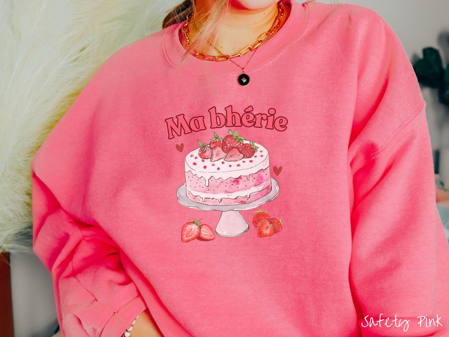 A Gildan sweatshirt featuring the phrase "Ma Bherie" is printed in bold red font above a charming illustration of a red-and-white strawberry cake displayed on a plate, topped with fresh strawberries and surrounded by even more berries.