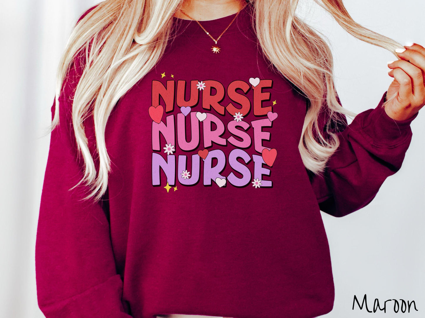 A stylish Gildan Sweatshirt featuring the word "NURSE" written three times vertically in red, pink, and purple font. There are red, white, and purple hearts mixed into the design as well as yellow twinkling stars.