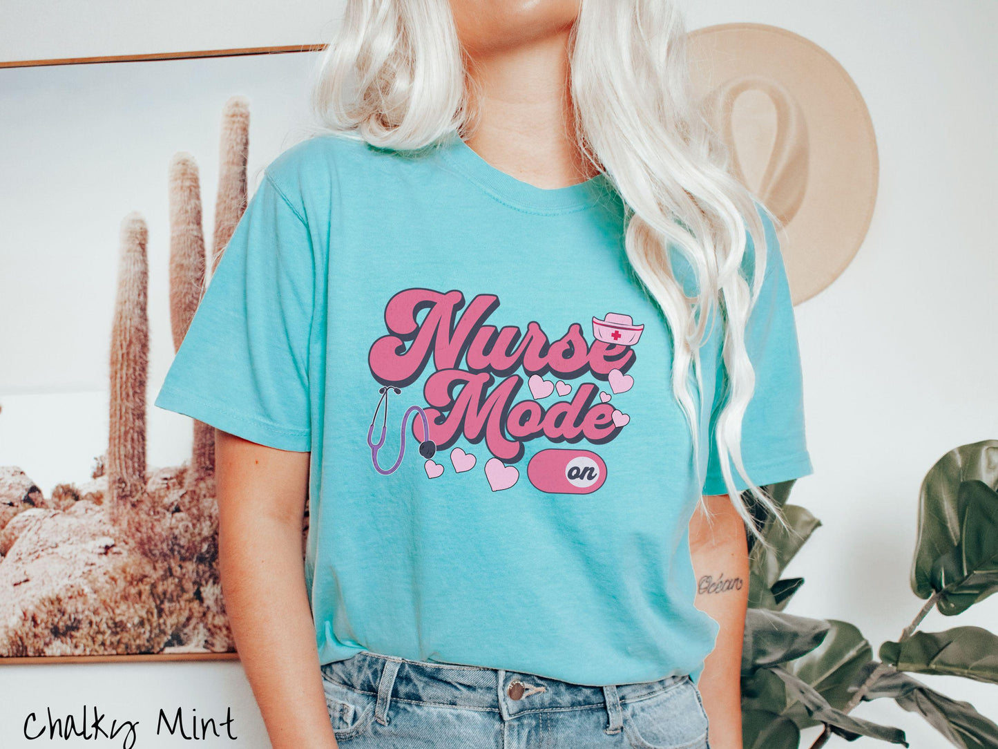 A stylish Comfort Colors t-shirt design features the text "Nurse Mode" in bold pink font with a pink nurse's cap placed over the letter E, and a stethoscope positioned beneath the text, alongside an "on" button widget set to the on position.