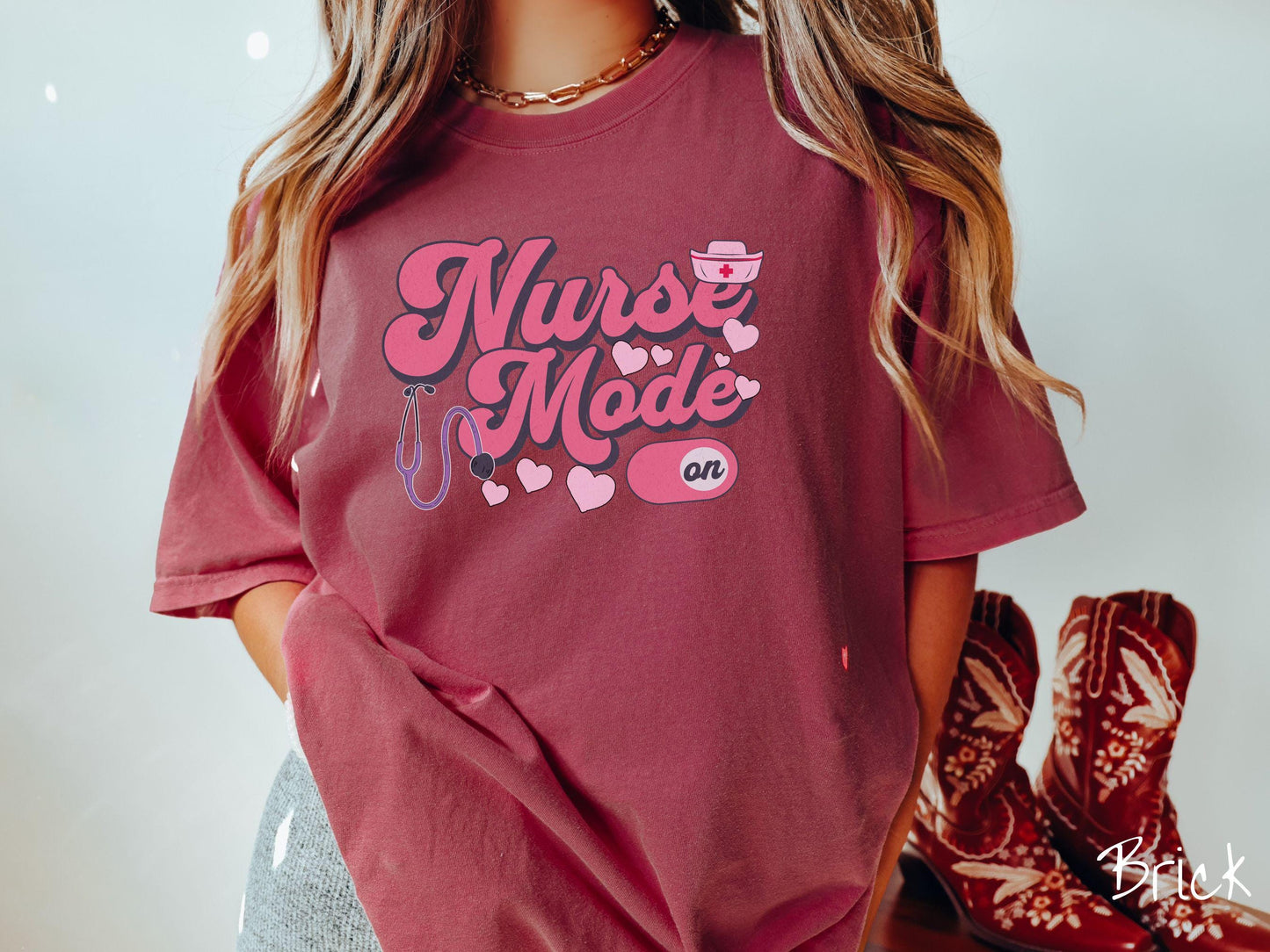 A stylish Comfort Colors t-shirt design features the text "Nurse Mode" in bold pink font with a pink nurse's cap placed over the letter E, and a stethoscope positioned beneath the text, alongside an "on" button widget set to the on position.