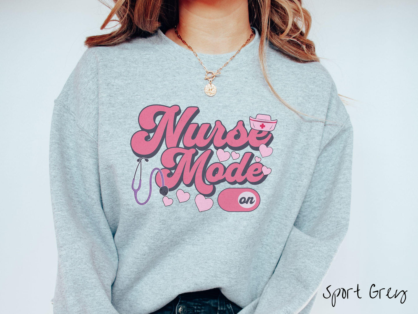 A stylish Gildan sweatshirt design features the text "Nurse Mode" in bold pink font with a pink nurse's cap placed over the letter E, and a stethoscope positioned beneath the text, alongside an "on" button widget set to the on position.