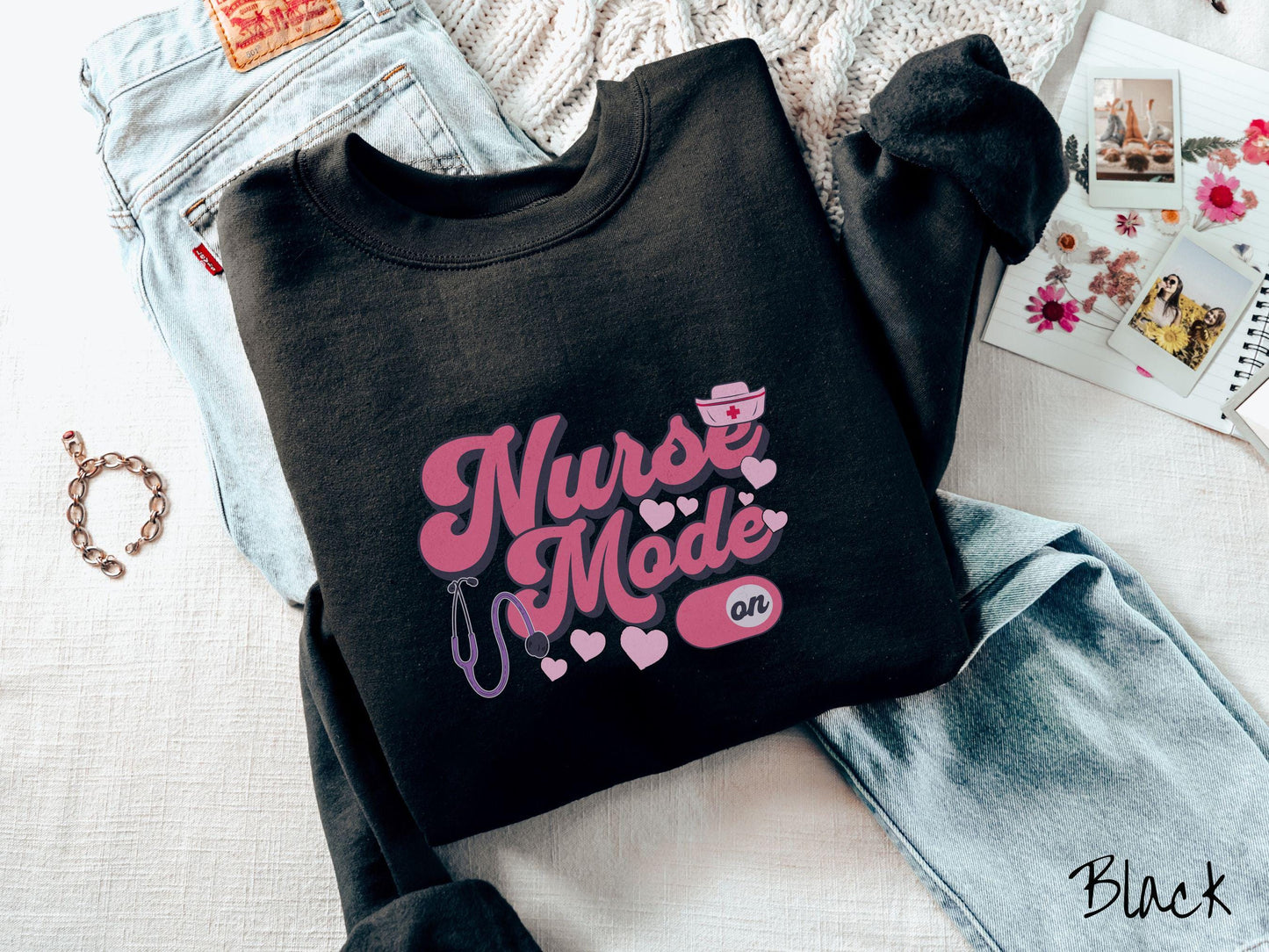 A stylish Gildan sweatshirt design features the text "Nurse Mode" in bold pink font with a pink nurse's cap placed over the letter E, and a stethoscope positioned beneath the text, alongside an "on" button widget set to the on position.