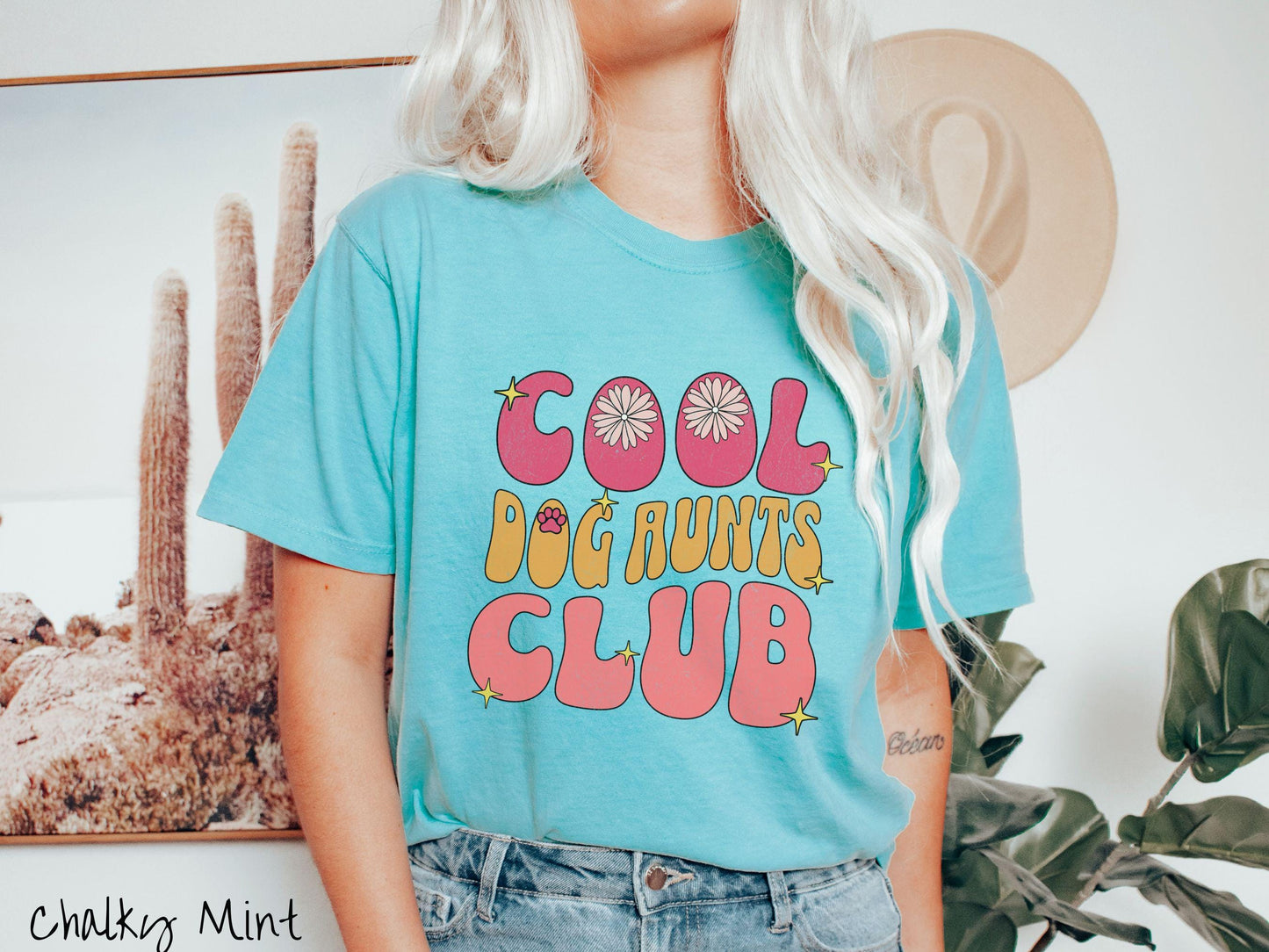A vintage Comfort Colors t-shirt featuring the text "Cool Dog Aunts Club" in bold red, yellow, and orange fonts with flower petals nestled in the "O" of COOL, a cute red paw print in the "O" of DOG, and sparkling yellow stars throughout.