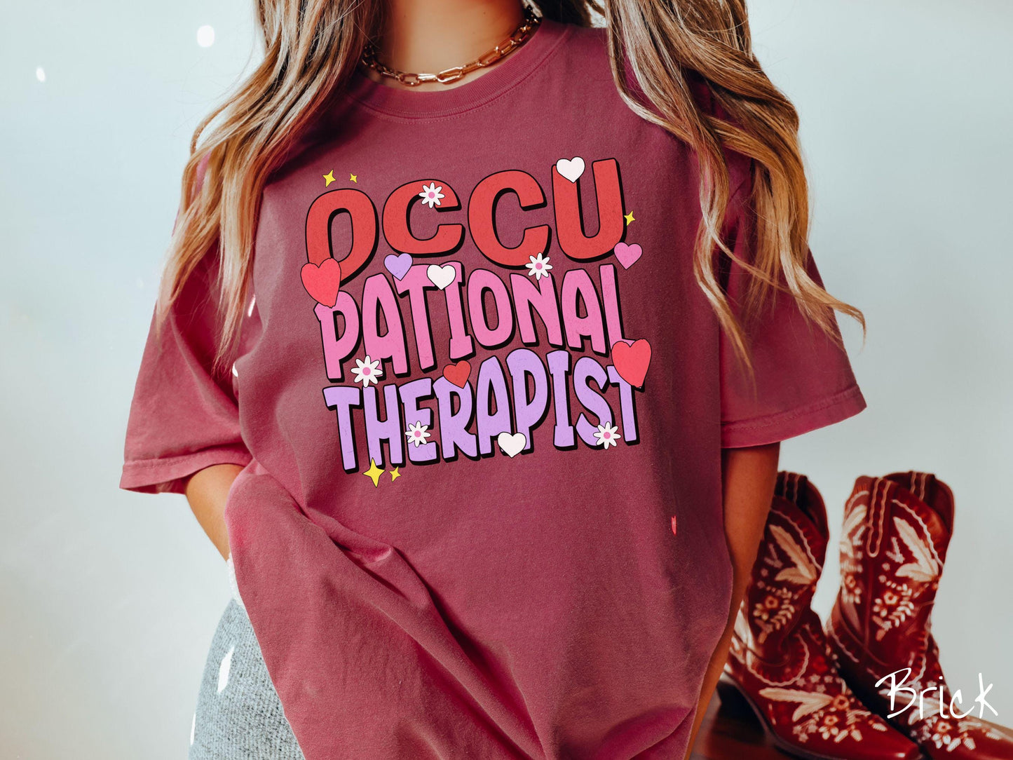 A stylish Comfort Colors t-shirt featuring the words "Occupational Therapist" arranged vertically in bold red, pink, and purple font, this design is enhanced with colorful hearts, flowers, and stars for a playful, uplifting touch.