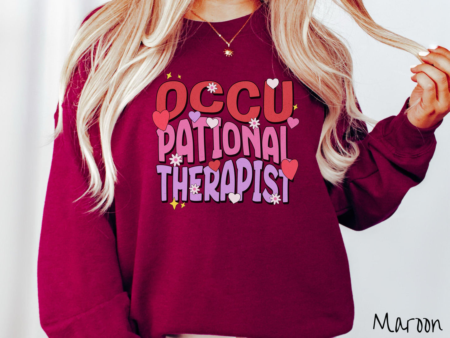 A stylish Gildan sweatshirt featuring the words "Occupational Therapist" arranged vertically in bold red, pink, and purple font, this design is enhanced with colorful hearts, flowers, and stars for a playful, uplifting touch.