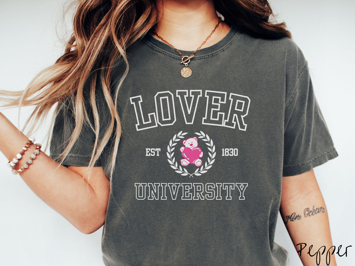 A vintage Comfort Colors t-shirt featuring a university logo-inspired design showcasing the text “Lover University” in collegiate-style font. Two pink laurel leaves frame a pink teddy bear hugging a heart with the text “EST. 1830” in pink font.