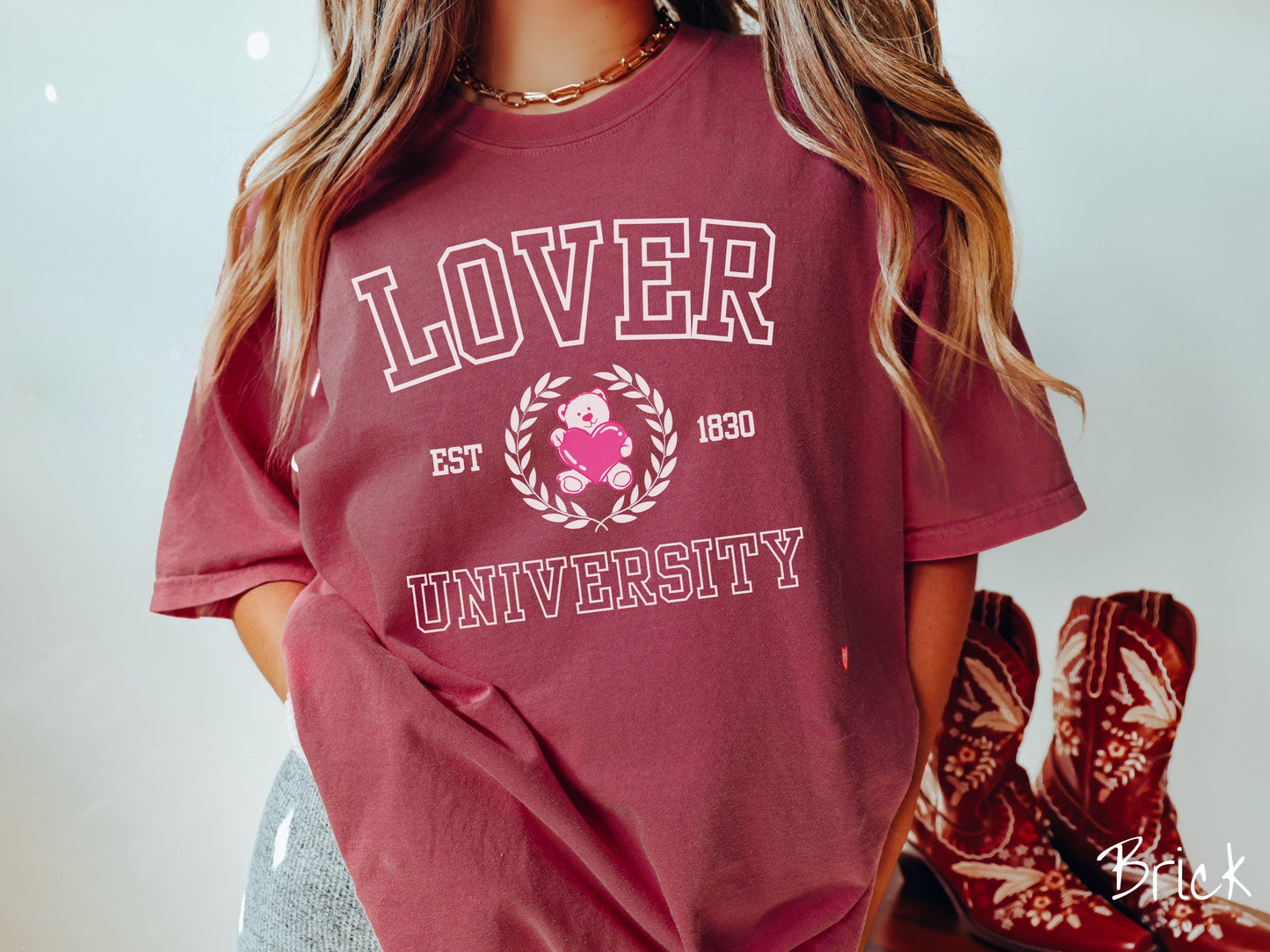 A vintage Comfort Colors t-shirt featuring a university logo-inspired design showcasing the text “Lover University” in collegiate-style font. Two pink laurel leaves frame a pink teddy bear hugging a heart with the text “EST. 1830” in pink font.