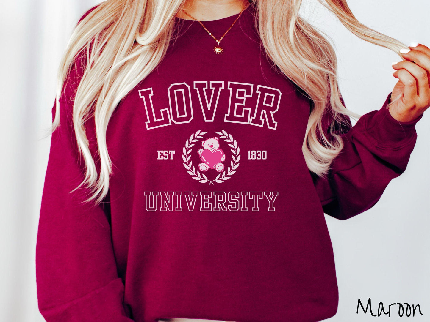 A vintage Gildan sweatshirt featuring a university logo-inspired design showcasing the text “Lover University” in collegiate-style font. Two pink laurel leaves frame a pink teddy bear hugging a heart with the text “EST. 1830” in pink font.