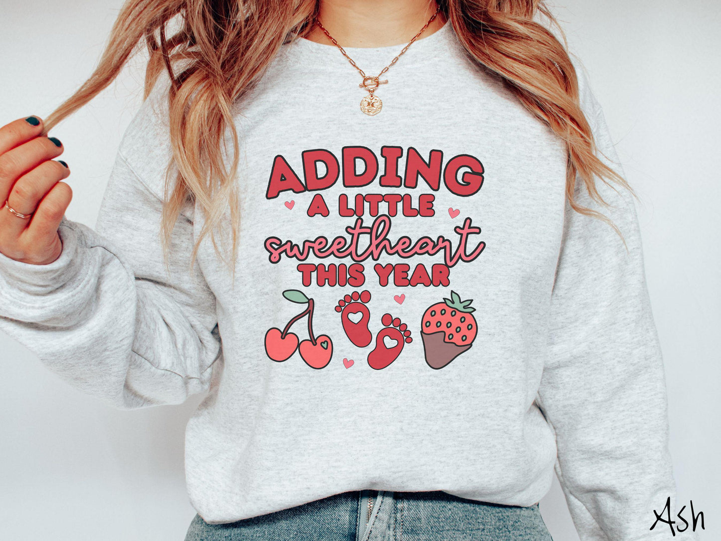 Share your exciting news with this vintage Gildan sweatshirt featuring the phrase “Adding a little sweetheart this year” in bold red font, paired with two red baby footprints framed by two red cherries and a chocolate-covered strawberry.