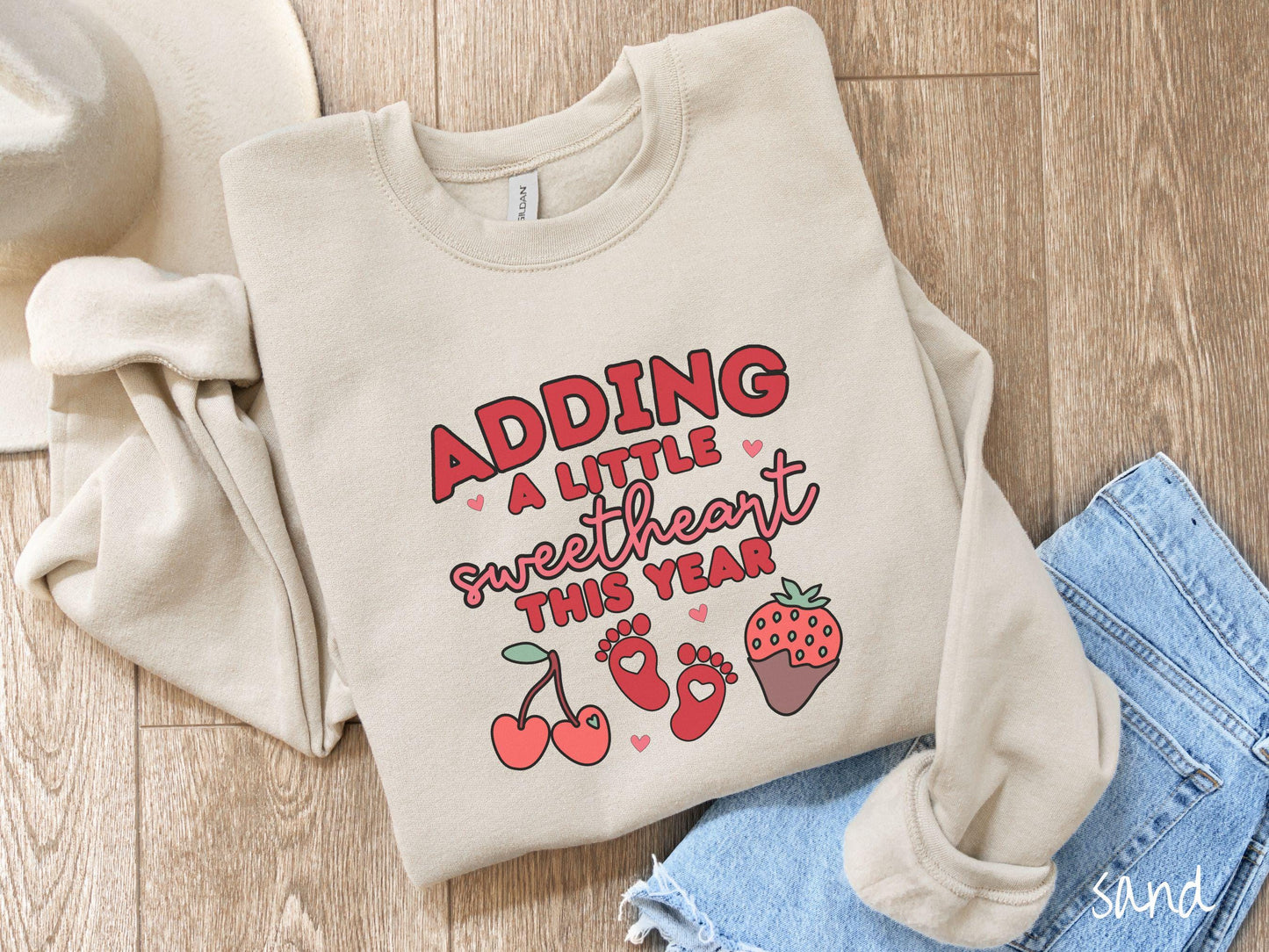 Share your exciting news with this vintage Gildan sweatshirt featuring the phrase “Adding a little sweetheart this year” in bold red font, paired with two red baby footprints framed by two red cherries and a chocolate-covered strawberry.