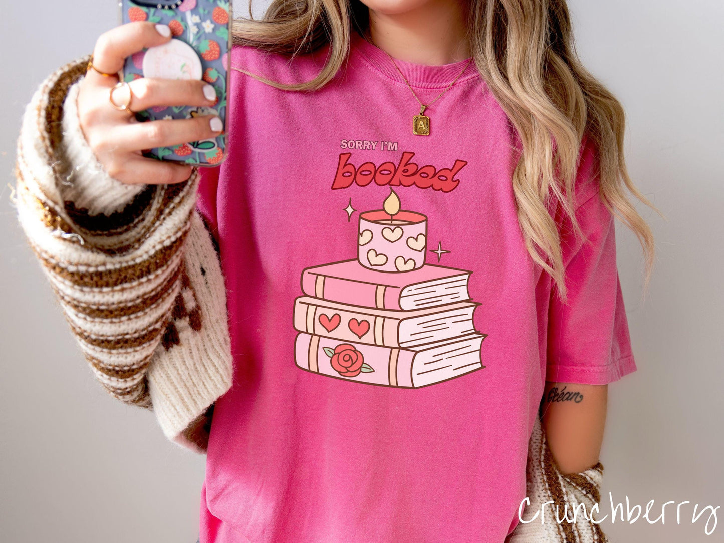 A vintage Comfort Colors t-shirt featuring the playful phrase “Sorry I’m Booked” in pink and red font, completed with a stack of three pink and purple books adorned with red roses and hearts on their spines, topped with a lit candle.