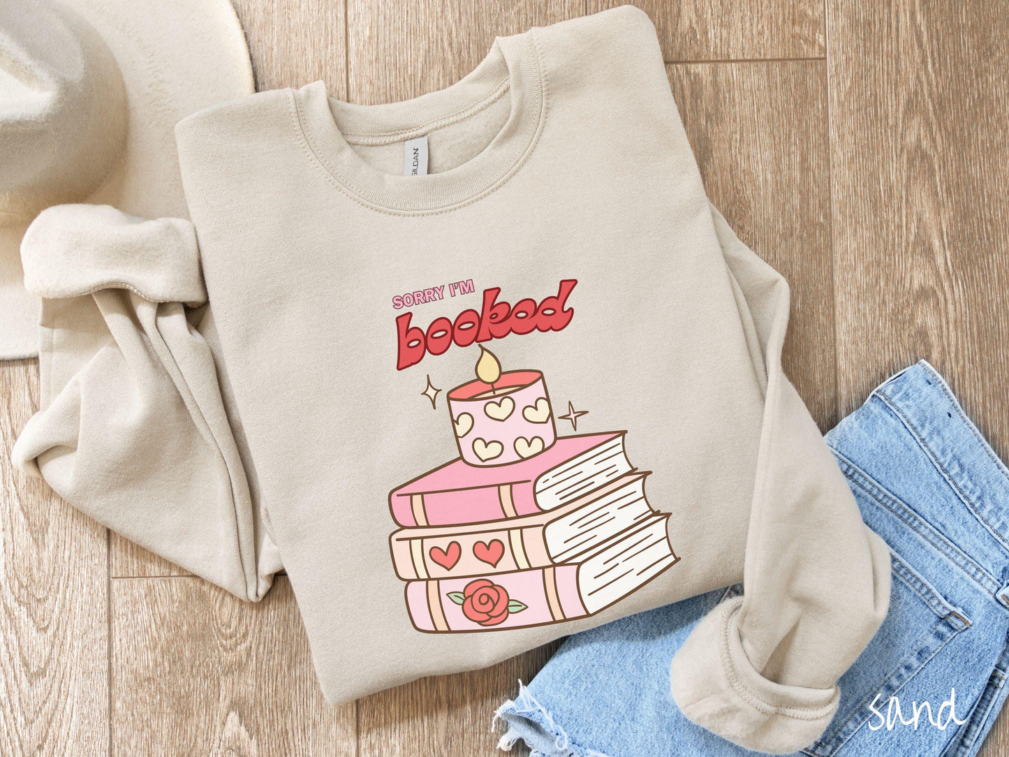A vintage Gildan sweatshirt featuring the playful phrase “Sorry I’m Booked” in pink and red font, completed with a stack of three pink and purple books adorned with red roses and hearts on their spines, topped with a lit candle.