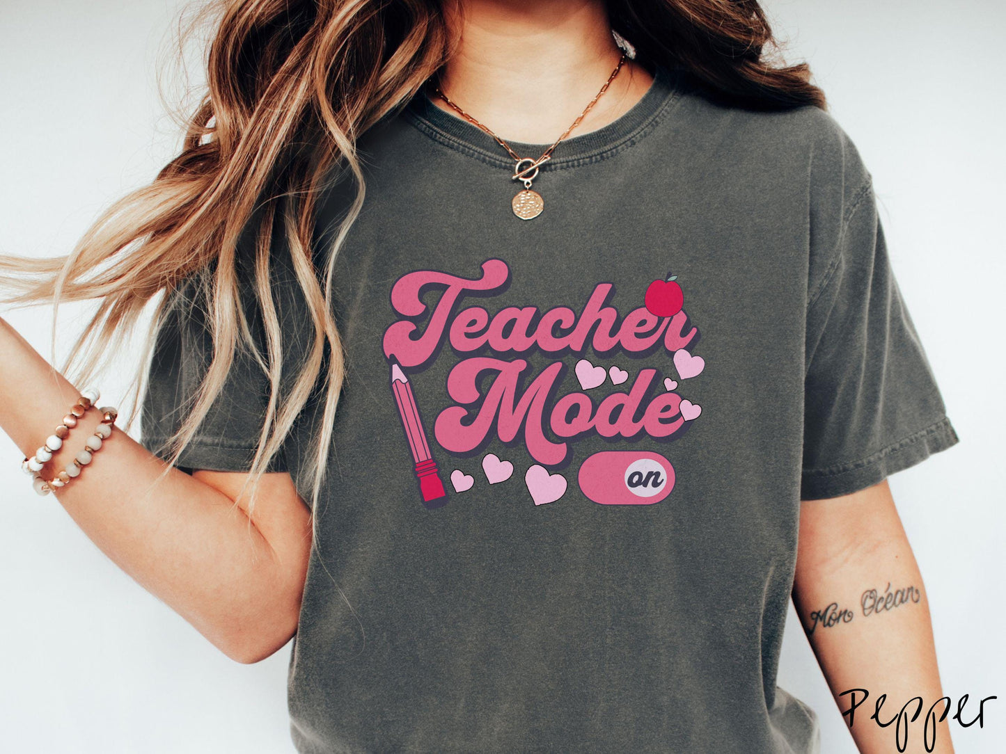 A vintage Comfort Colors t-shirt featuring the phrase “Teacher Mode” in playful pink font, an “On” button toggled to the “On” position, and with charming details like a red pencil, a red apple, and pink hearts sprinkled throughout.