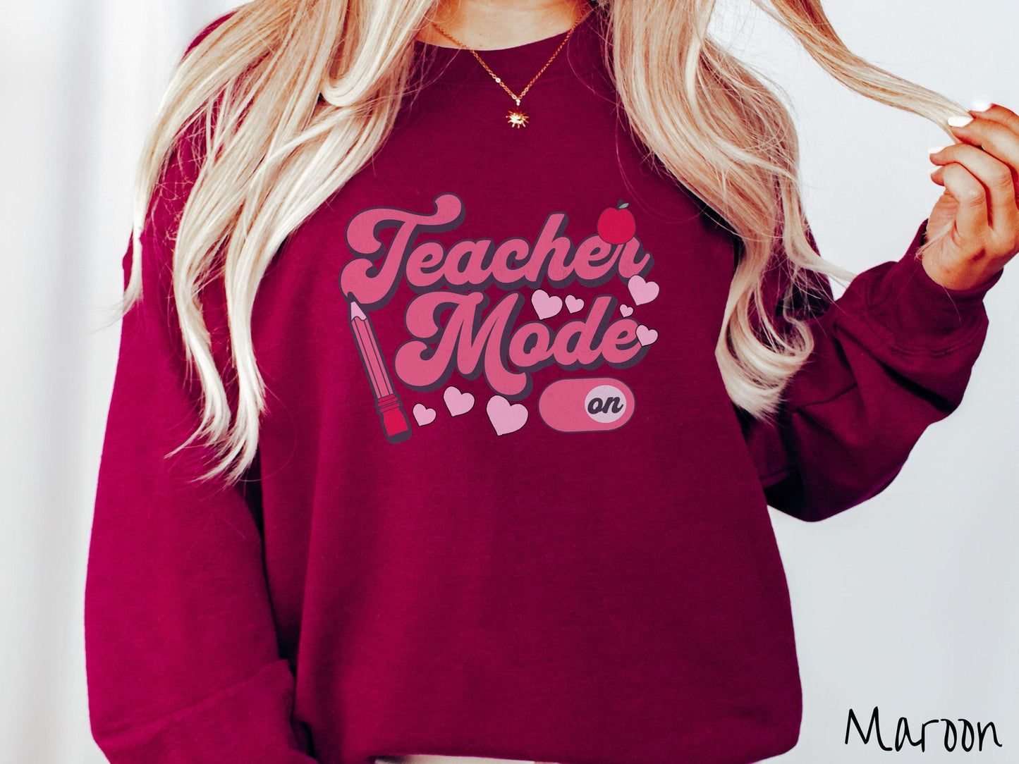 A vintage Gildan sweathirt featuring the phrase “Teacher Mode” in playful pink font, an “On” button toggled to the “On” position, and with charming details like a red pencil, a red apple, and pink hearts sprinkled throughout.