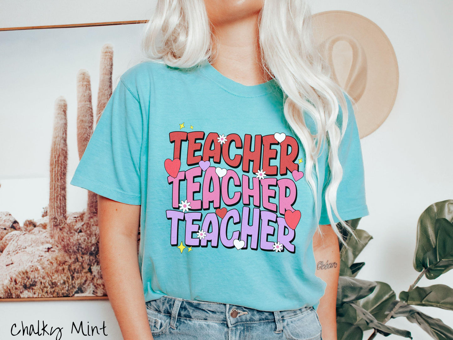 A vibrant vintage Comfort Colors t-shirt featuring the word “Teacher” written vertically three times in bold red, pink, and purple font, with colorful hearts, flowers, and twinkling stars scattered throughout for a whimsical and eye-catching look.