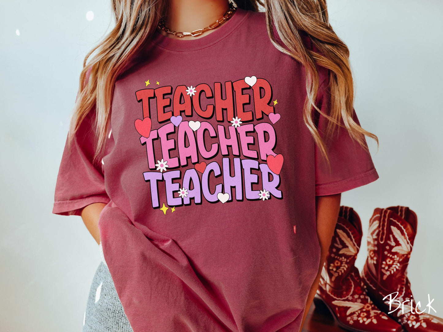 A vibrant vintage Comfort Colors t-shirt featuring the word “Teacher” written vertically three times in bold red, pink, and purple font, with colorful hearts, flowers, and twinkling stars scattered throughout for a whimsical and eye-catching look.