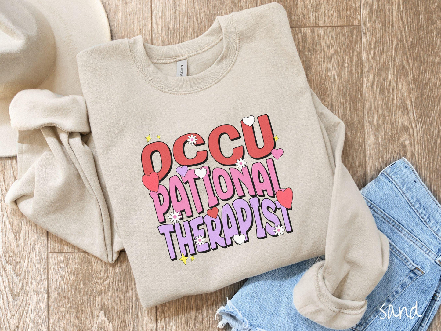 A stylish Gildan sweatshirt featuring the words "Occupational Therapist" arranged vertically in bold red, pink, and purple font, this design is enhanced with colorful hearts, flowers, and stars for a playful, uplifting touch.