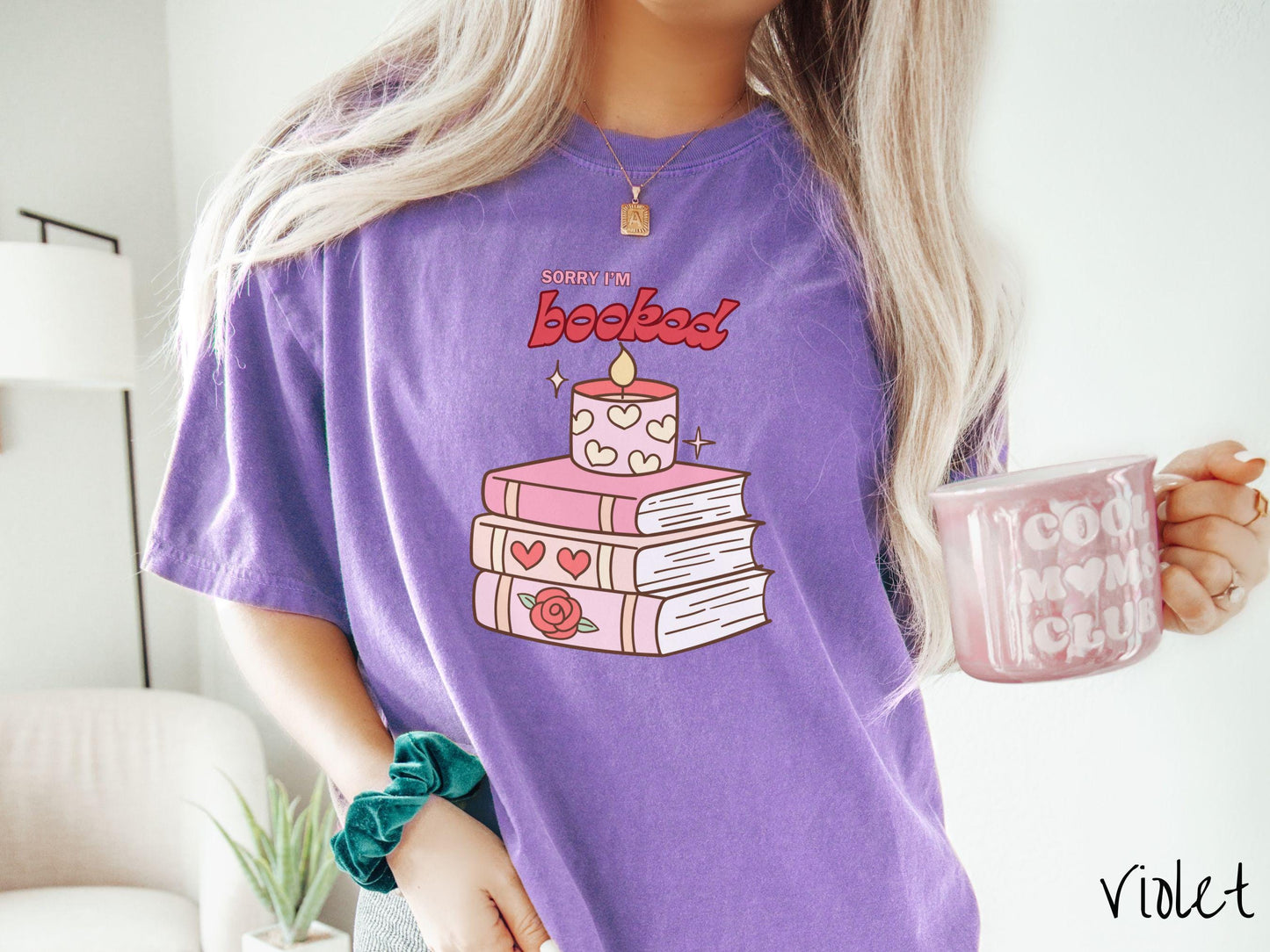 A vintage Comfort Colors t-shirt featuring the playful phrase “Sorry I’m Booked” in pink and red font, completed with a stack of three pink and purple books adorned with red roses and hearts on their spines, topped with a lit candle.