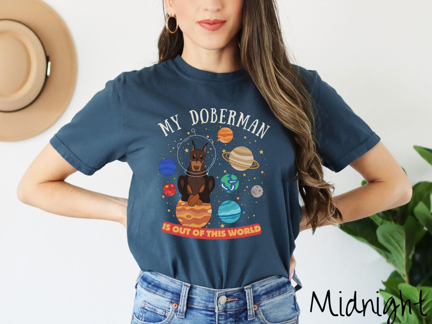A vintage Comfort Colors t-shirt featuring the text My Doberman is Out of This World. The design showcases a Doberman sitting proudly atop Jupiter, surrounded by a vibrant solar system filled with colorful planets and twinkling stars.