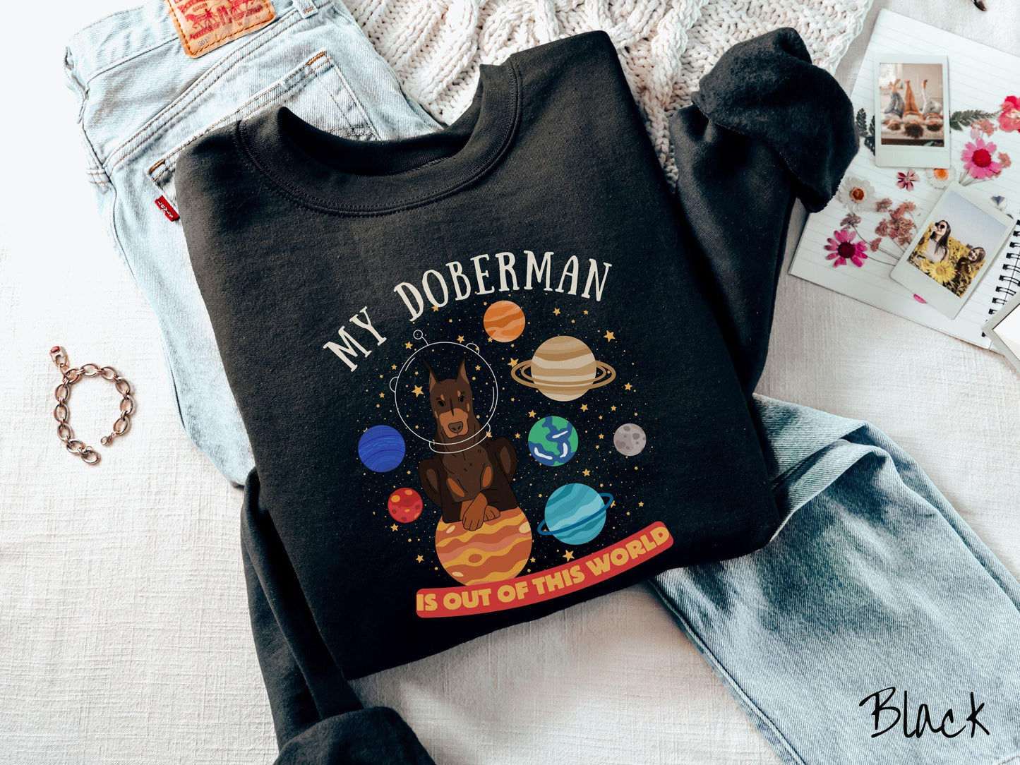 A vintage Gildan sweatshirt featuring the text My Doberman is Out of This World. The design showcases a Doberman sitting proudly atop Jupiter, surrounded by a vibrant solar system filled with colorful planets and twinkling stars.