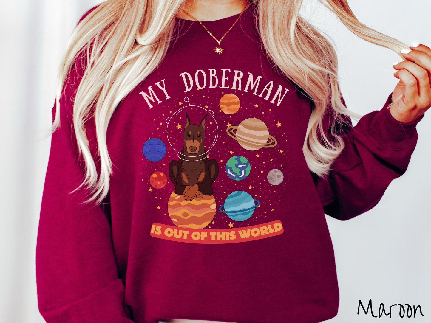 A vintage Gildan sweatshirt featuring the text My Doberman is Out of This World. The design showcases a Doberman sitting proudly atop Jupiter, surrounded by a vibrant solar system filled with colorful planets and twinkling stars.