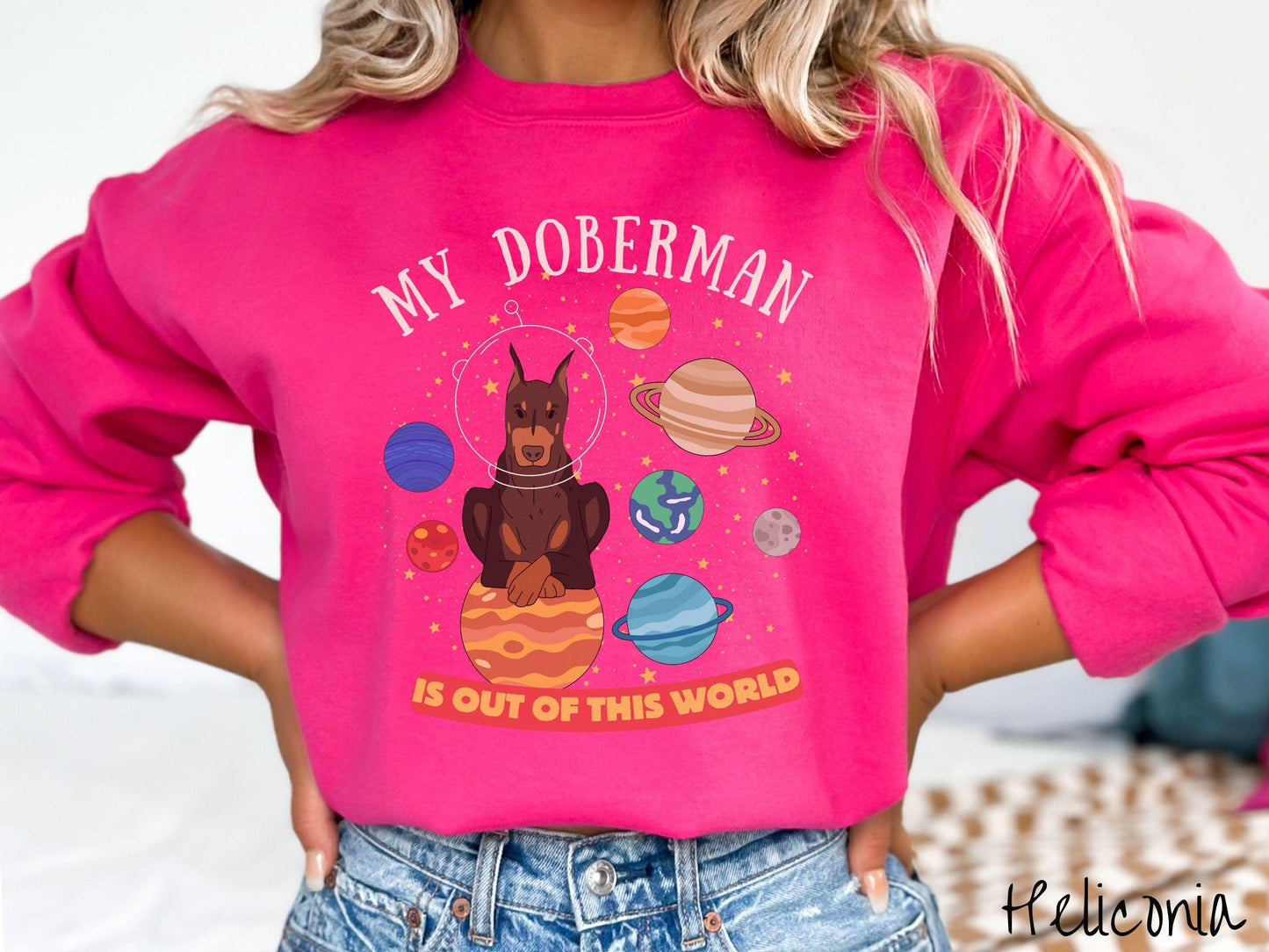 A vintage Gildan sweatshirt featuring the text My Doberman is Out of This World. The design showcases a Doberman sitting proudly atop Jupiter, surrounded by a vibrant solar system filled with colorful planets and twinkling stars.