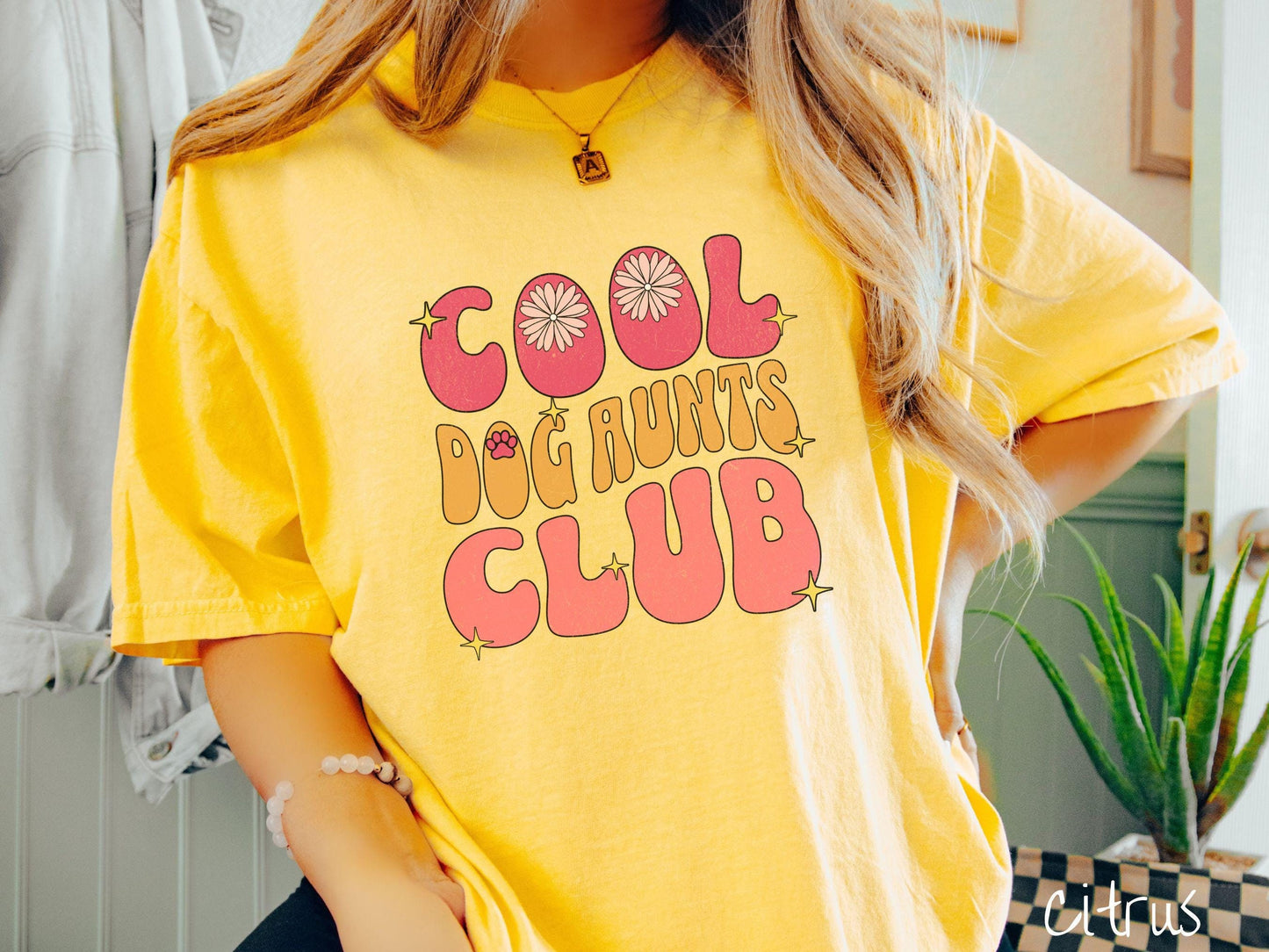 A vintage Comfort Colors t-shirt featuring the text "Cool Dog Aunts Club" in bold red, yellow, and orange fonts with flower petals nestled in the "O" of COOL, a cute red paw print in the "O" of DOG, and sparkling yellow stars throughout.