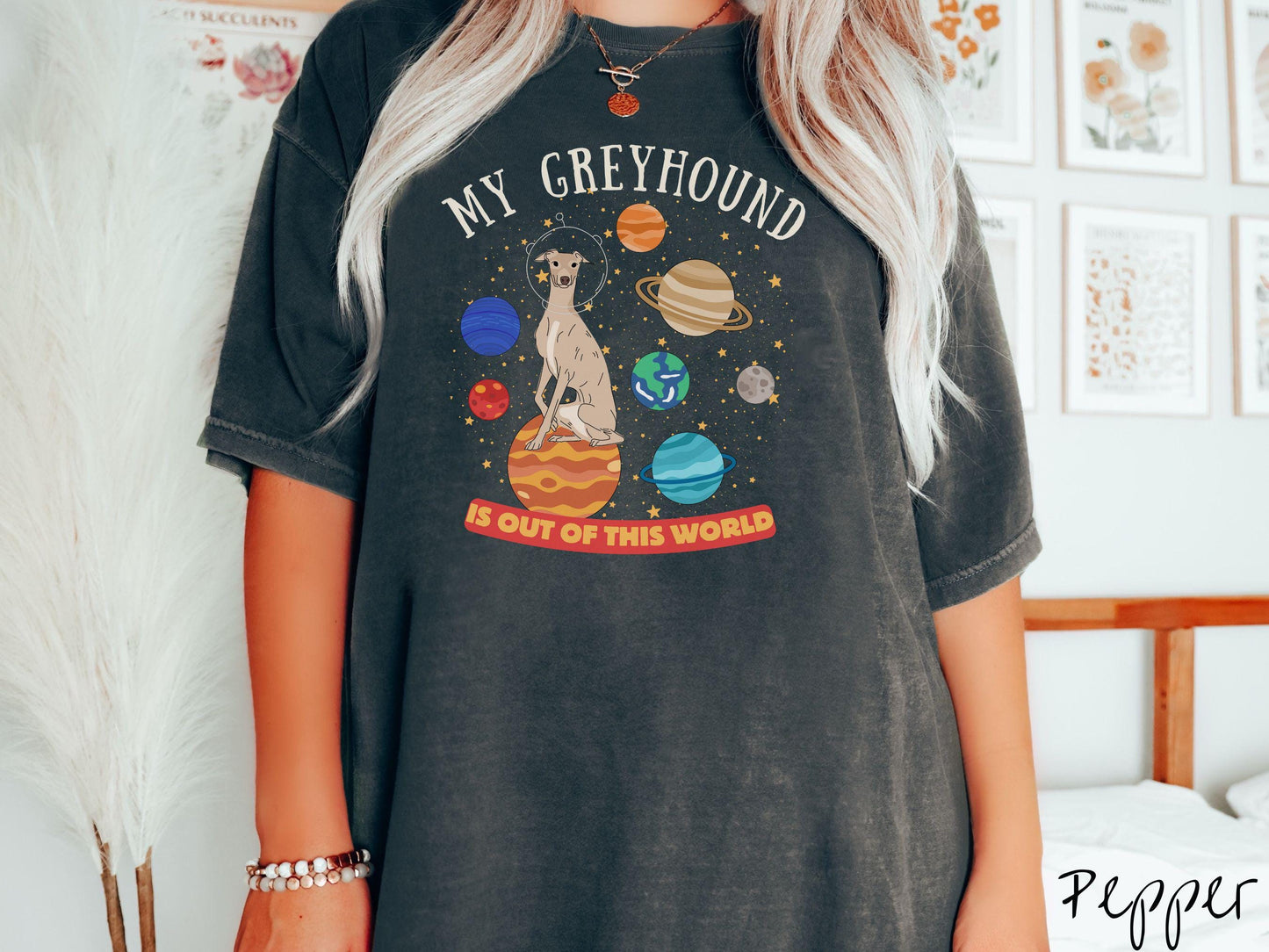 A vintage Comfort Colors t-shirt featuring the text My Greyhound is Out of This World. The design showcases a Greyhound sitting proudly atop Jupiter, surrounded by a vibrant solar system filled with colorful planets and twinkling stars.