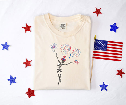 A woman wearing a cute, vintage ivory colored Comfort Colors t-shirt with a skeleton wearing a red, white, and blue patriotic top hat dancing under fireworks holding an American flag.