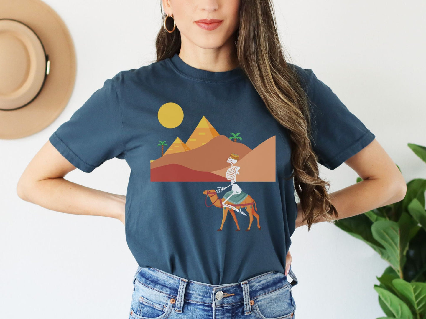 A woman wearing a cute, vintage navy blue colored shirt with a skeleton wearing an Egyptian hat riding a brown camel through the desert, the Egyptian pyramids in the distance behind sand dunes and palm trees with the sun high in the sky.